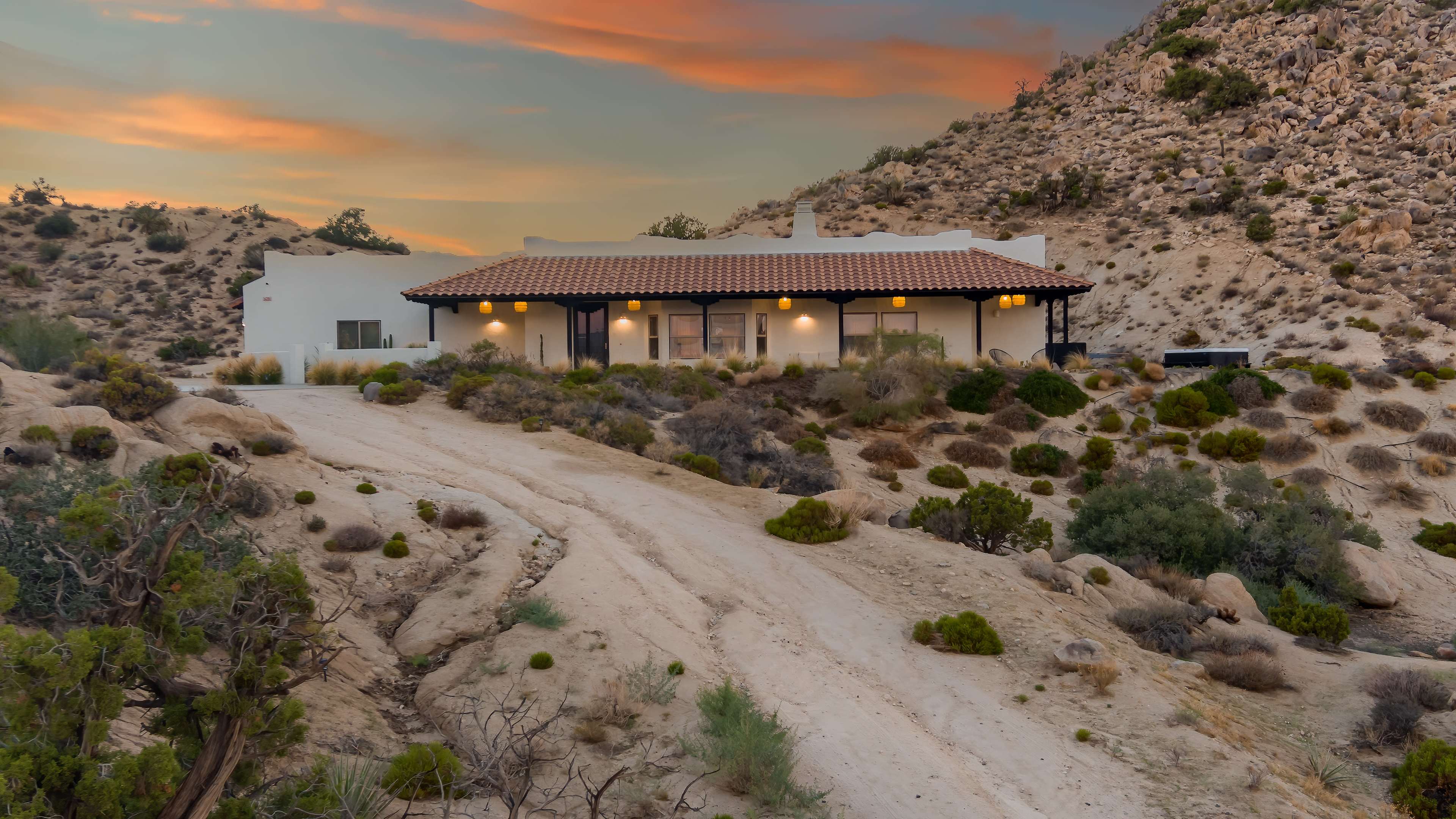 A single-story white adobe house with a terracotta roof is situated on a sandy, uneven landscape surrounded by desert vegetation and rocky hills under a colorful sunset sky.