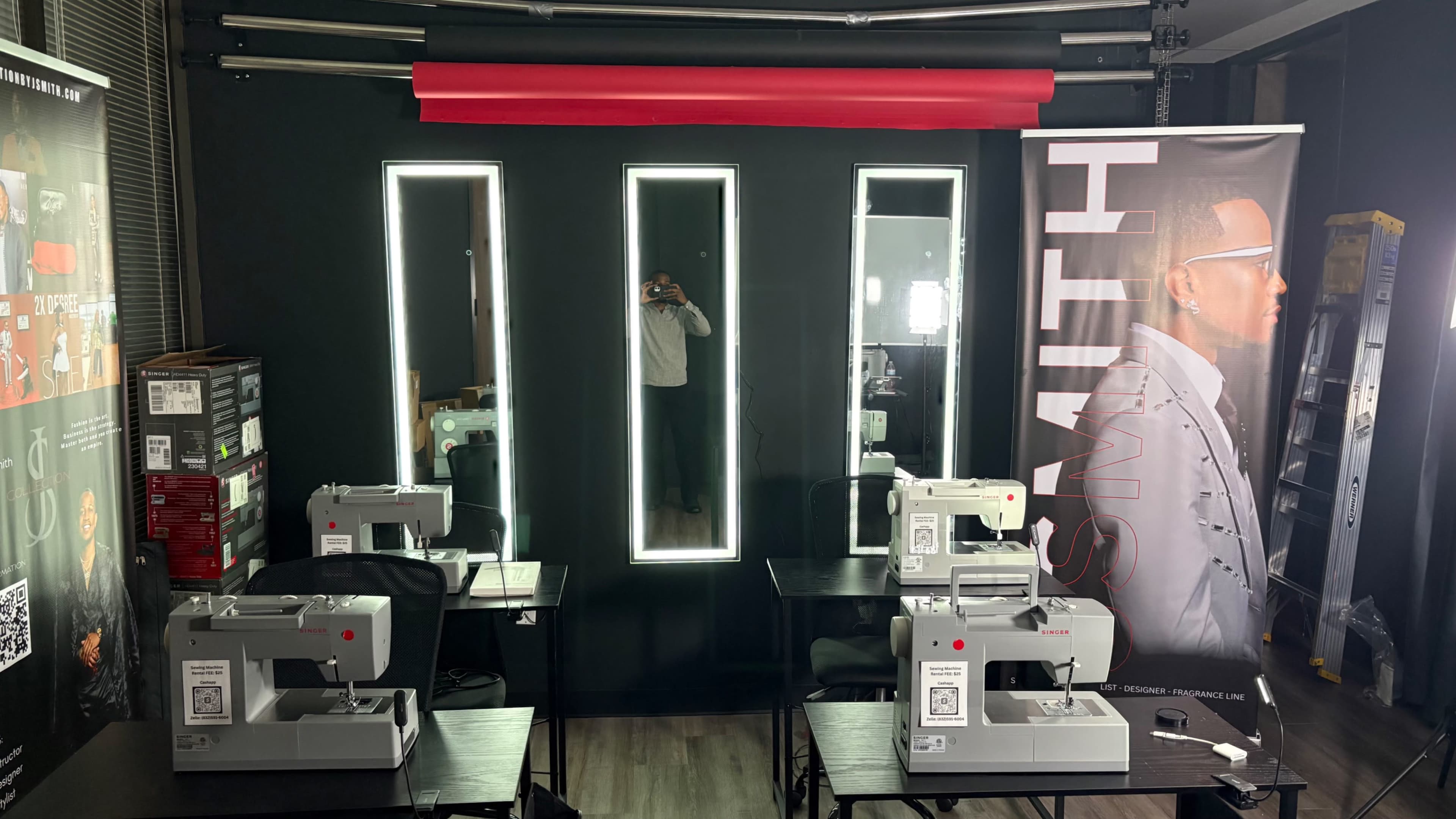 The image shows a sewing studio with several sewing machines arranged on tables, illuminated by vertical mirrors and a banner featuring a model.