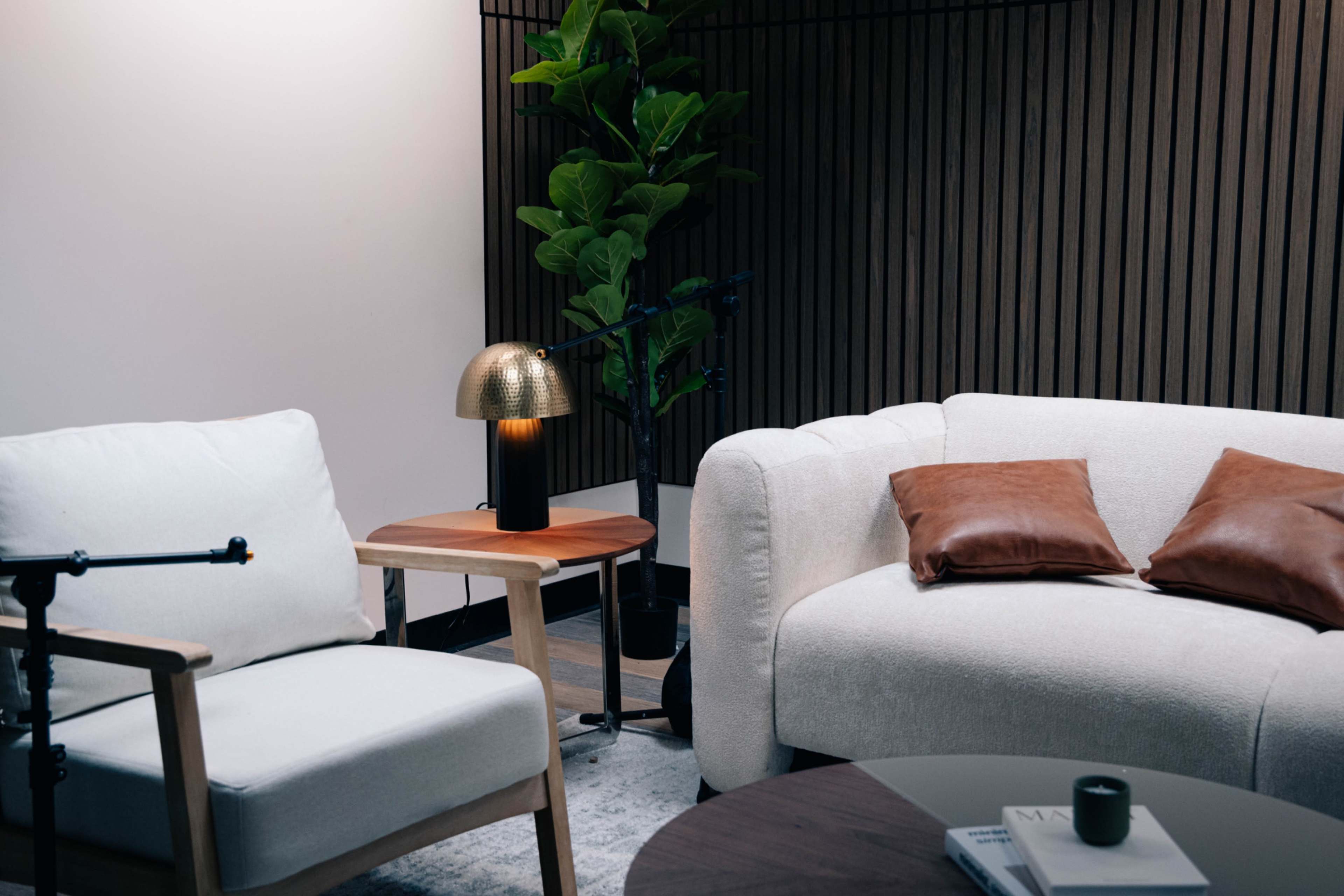 A cozy sitting area features a white sofa with brown cushions, a light-colored armchair, a circular coffee table, and a lamp on a side table, with a backdrop of wooden paneling and a potted plant.