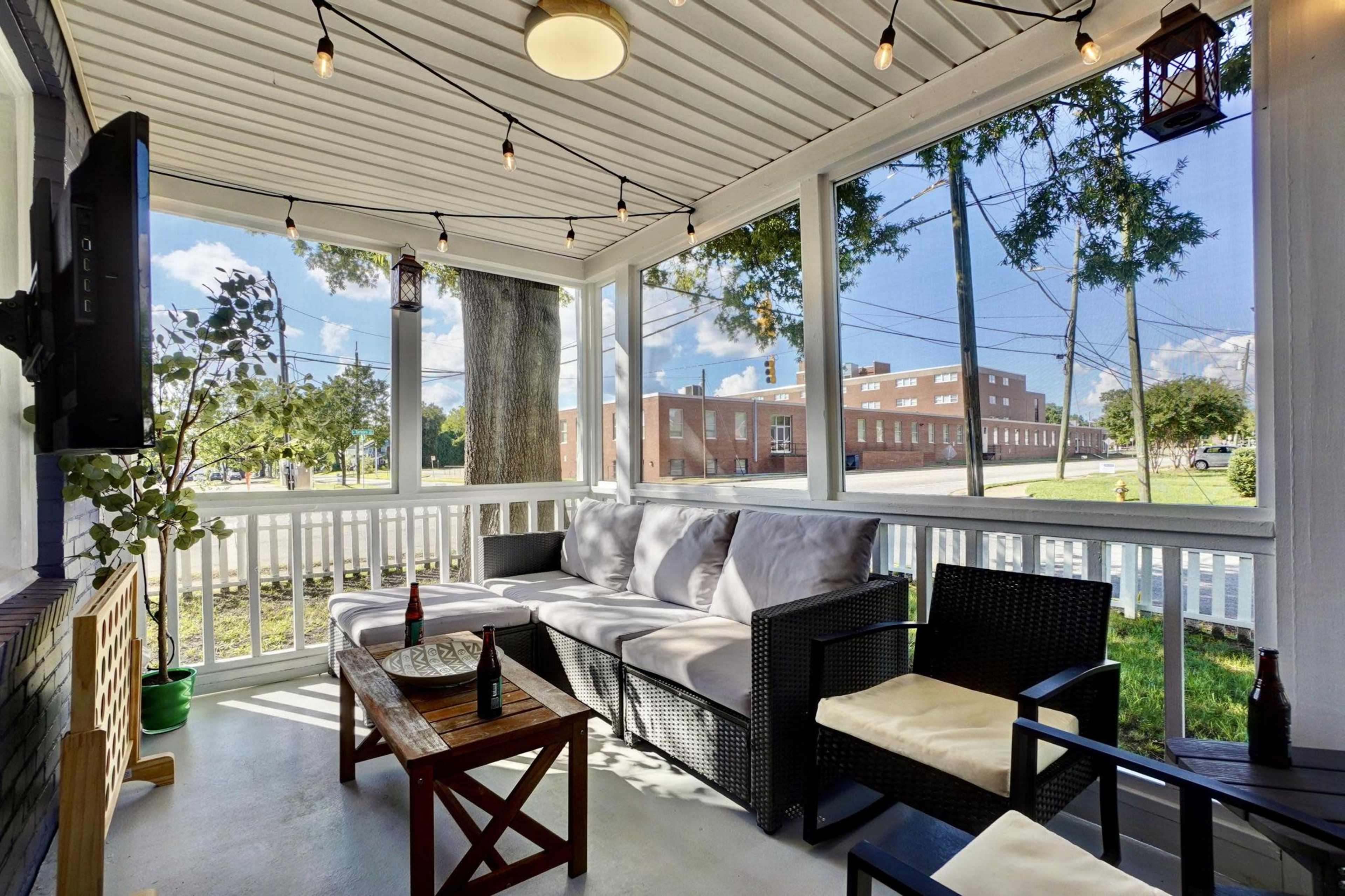 A screened porch features a gray sofa, wooden coffee table, and several chairs, illuminated by string lights, with a view of a nearby building and trees outside.