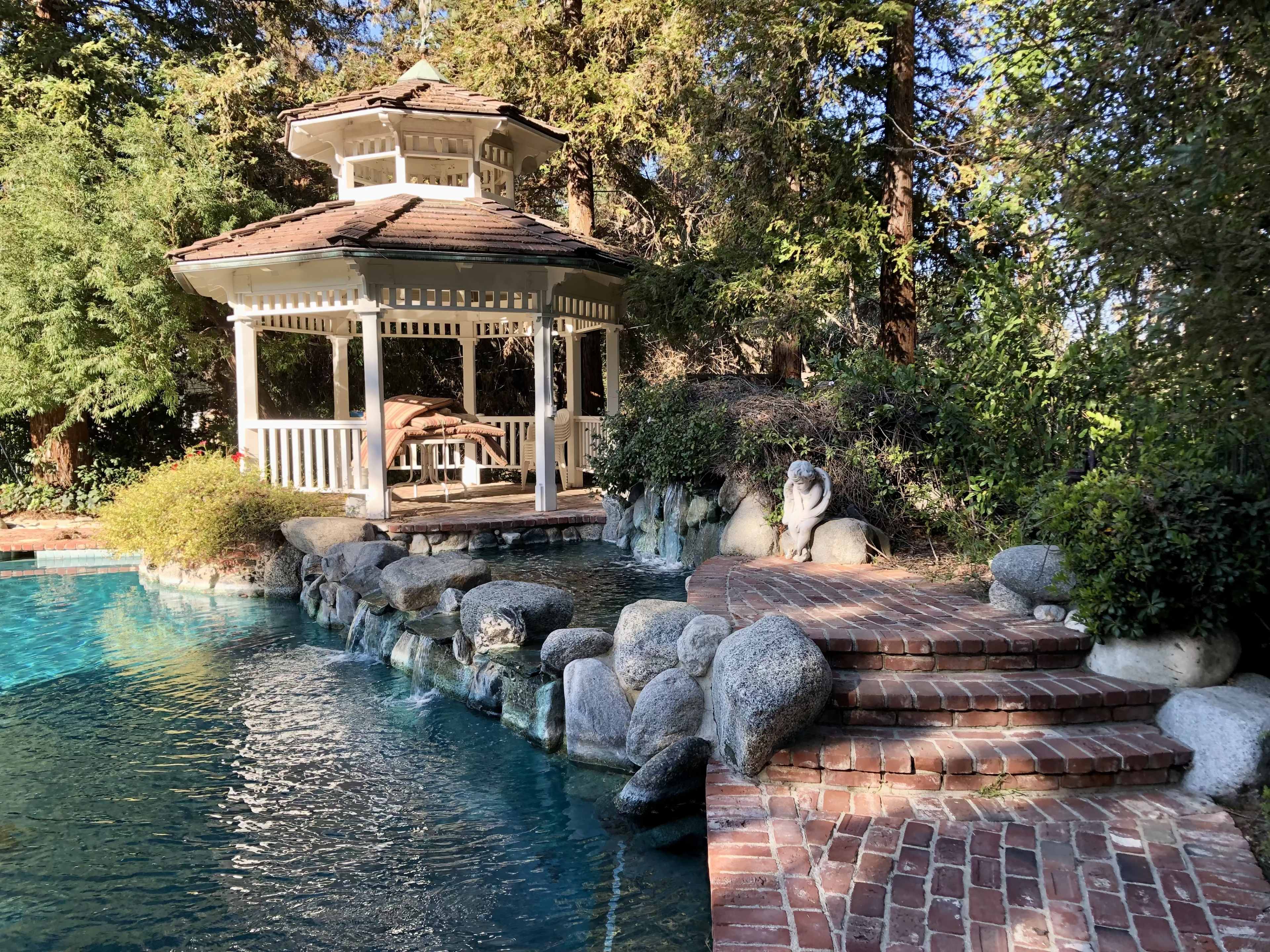 A gazebo stands beside a stone-lined swimming pool with a brick pathway leading to it, surrounded by greenery.