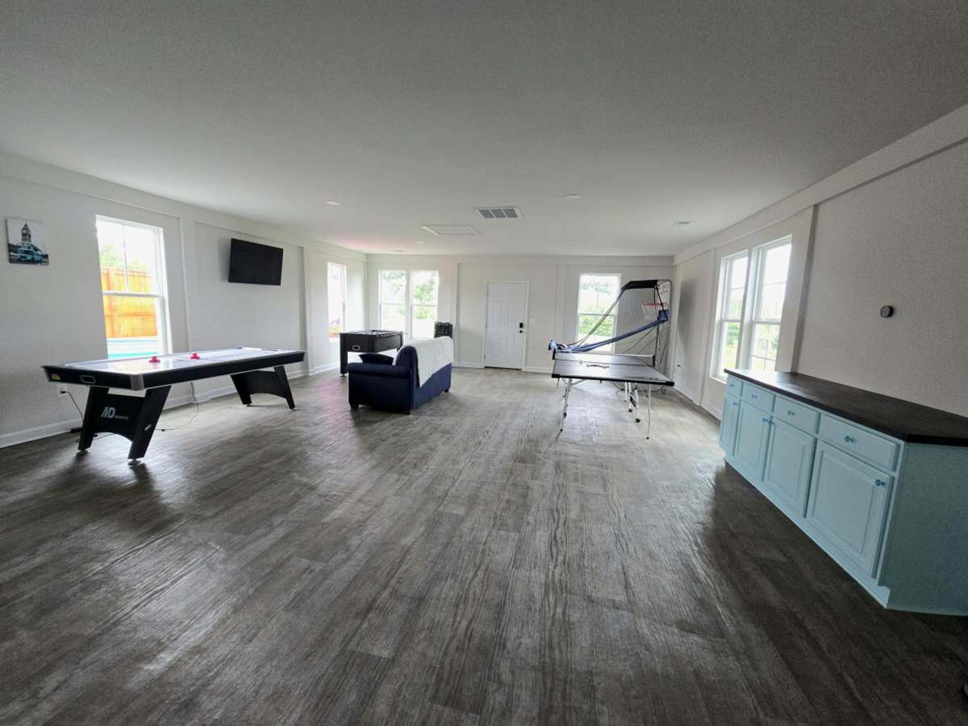 The image shows a spacious indoor recreation room featuring a pool table, a table tennis setup, a couch, and multiple windows allowing natural light.