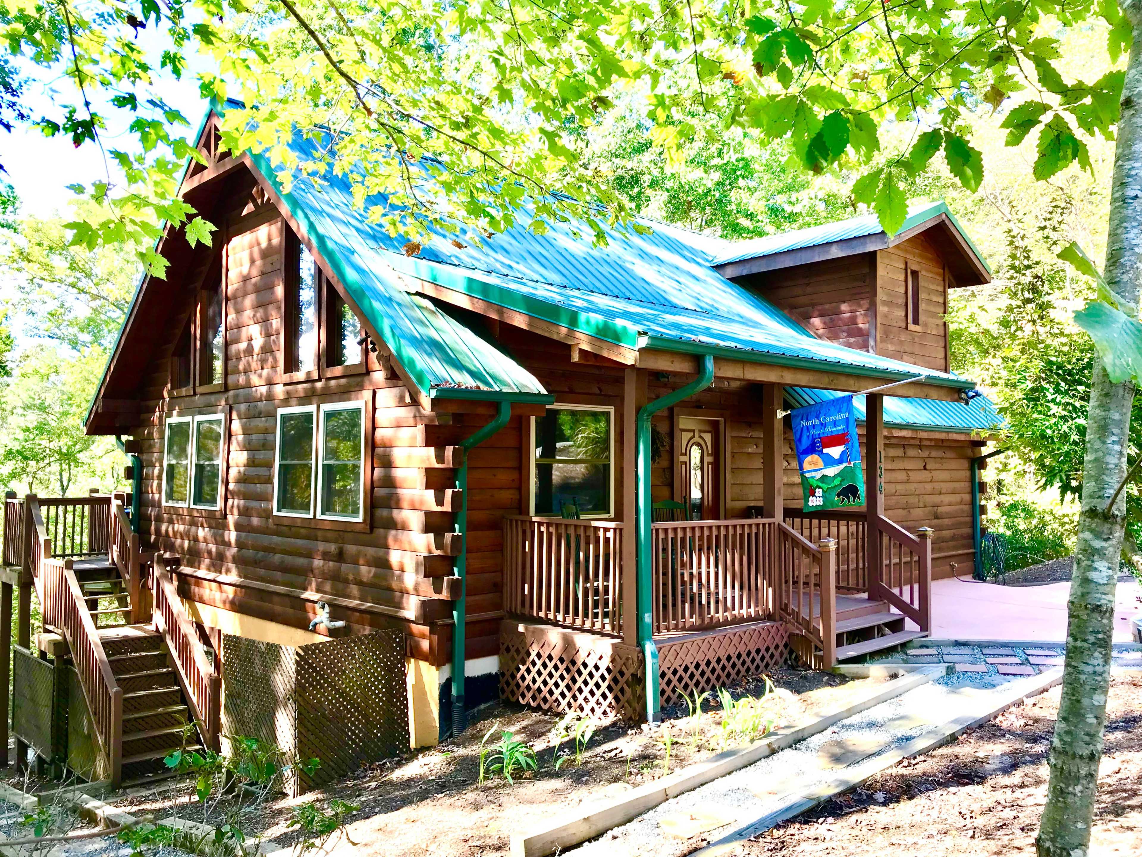 A wooden cabin with a green metal roof features large windows and a covered porch surrounded by trees.