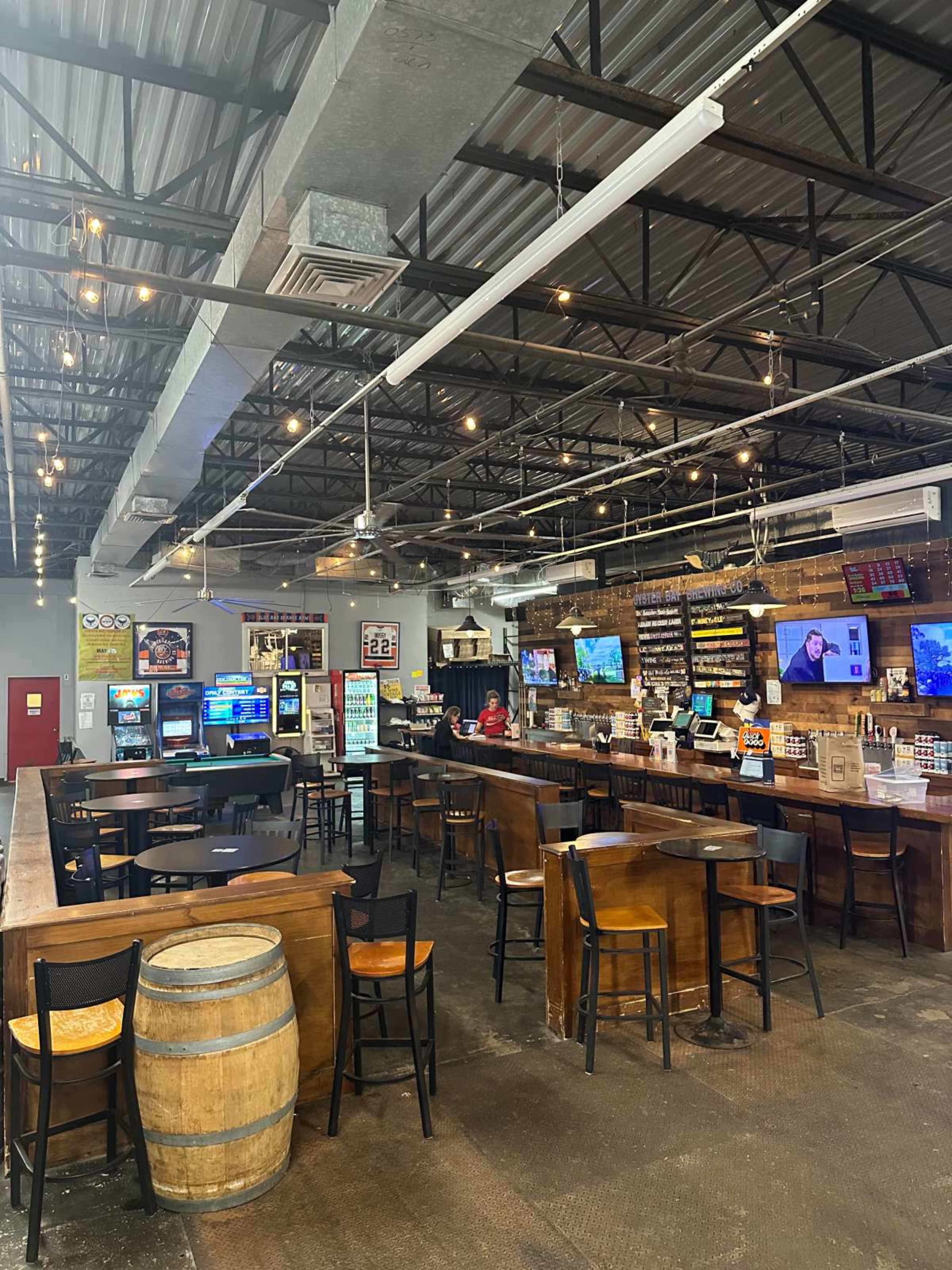 The image shows a spacious bar interior with wooden tables and chairs, a long counter, and multiple screens displaying various content.