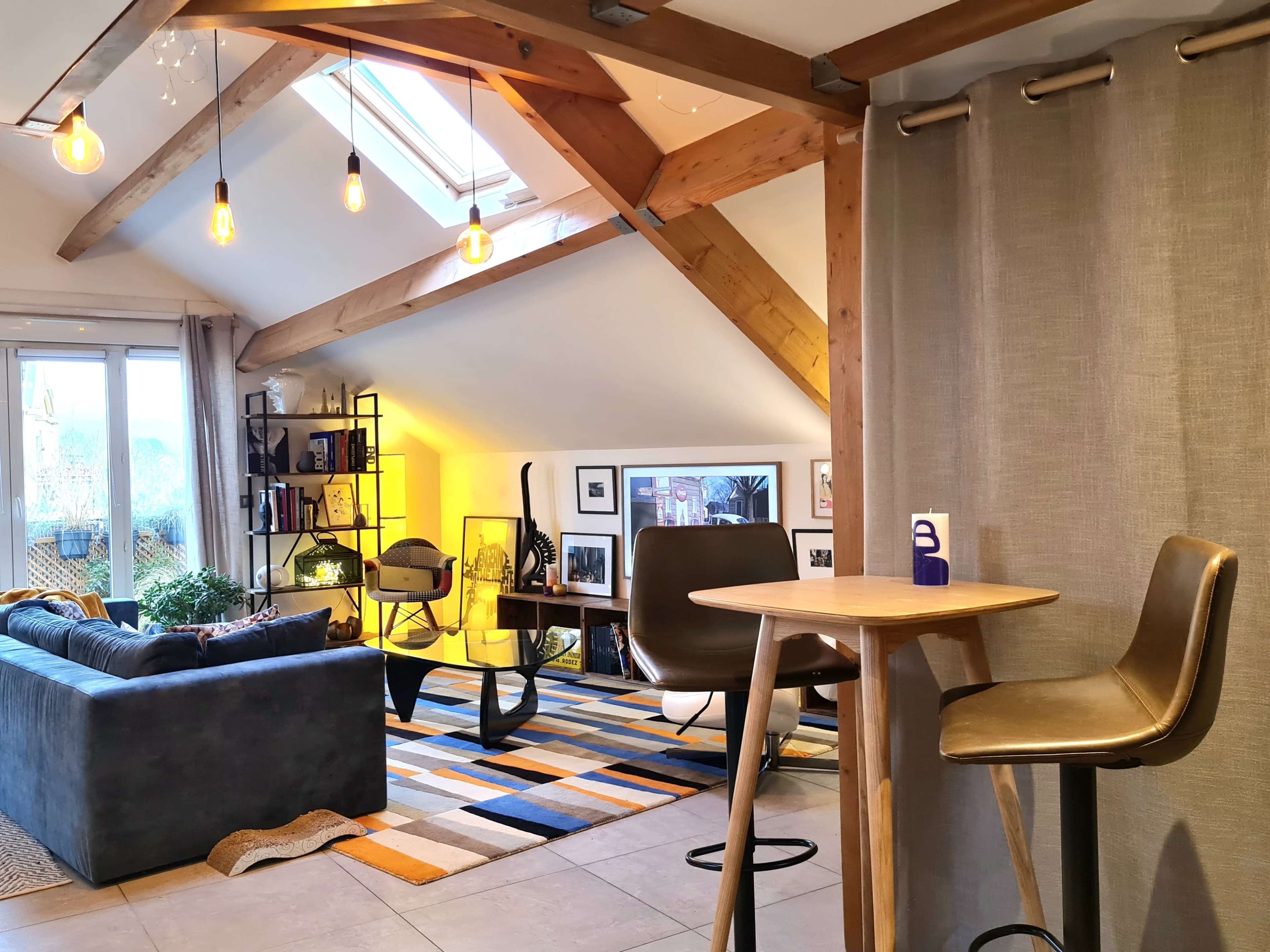 A modern living space with a high ceiling, featuring a sofa, a small table with stools, and a bookshelf illuminated by warm light.
