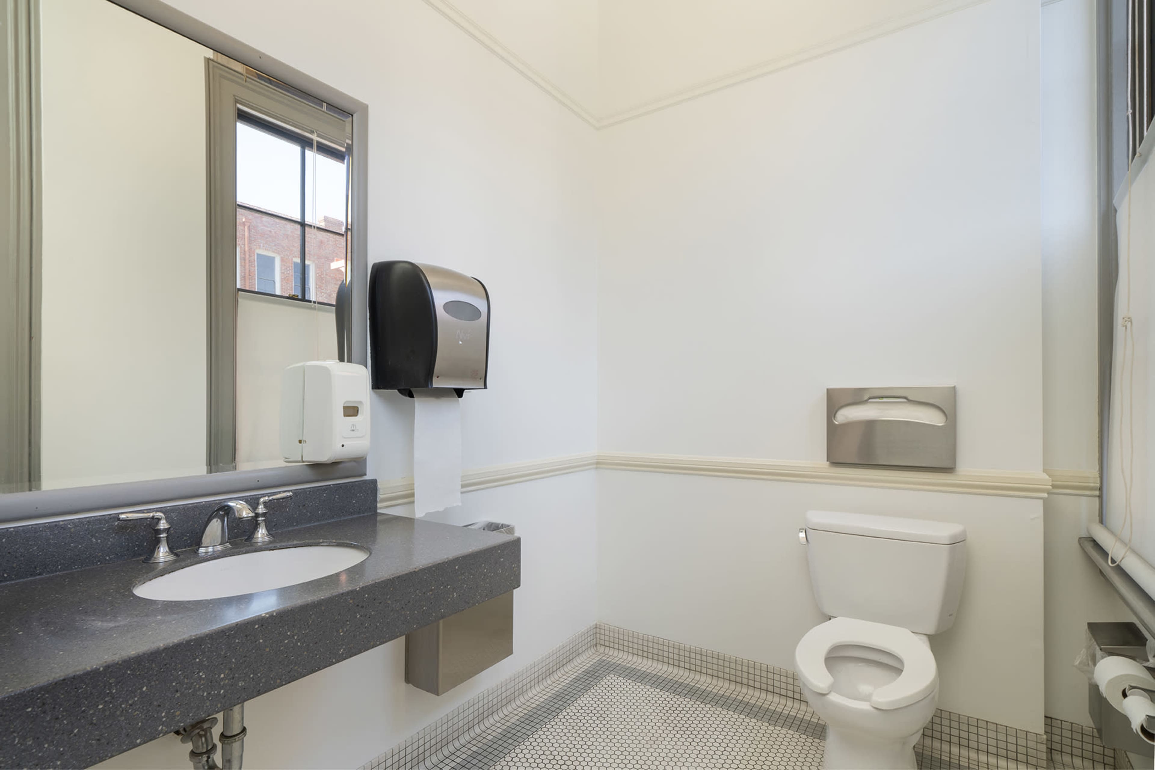 A clean restroom featuring a toilet, a sink with two faucets, and a paper towel dispenser on the wall.