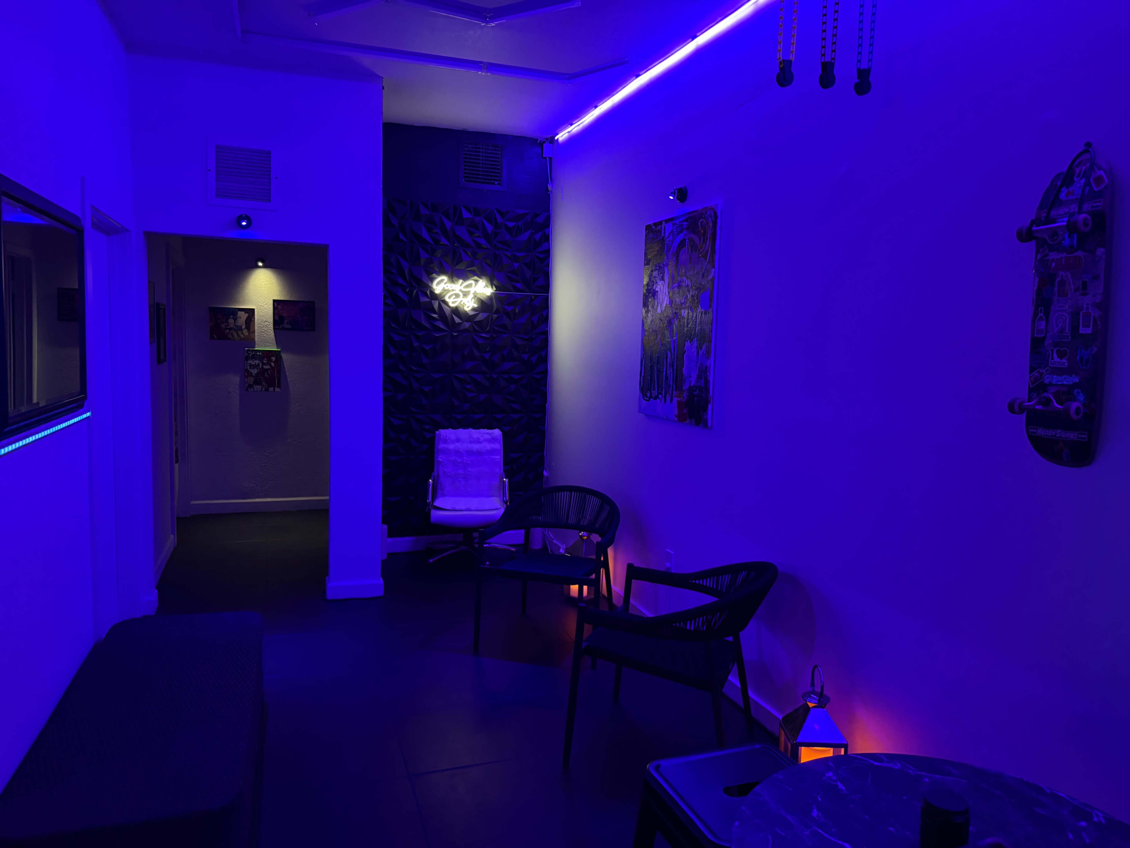 The image shows a dimly lit room with purple lighting, featuring a couple of black chairs, a small table, and wall art.
