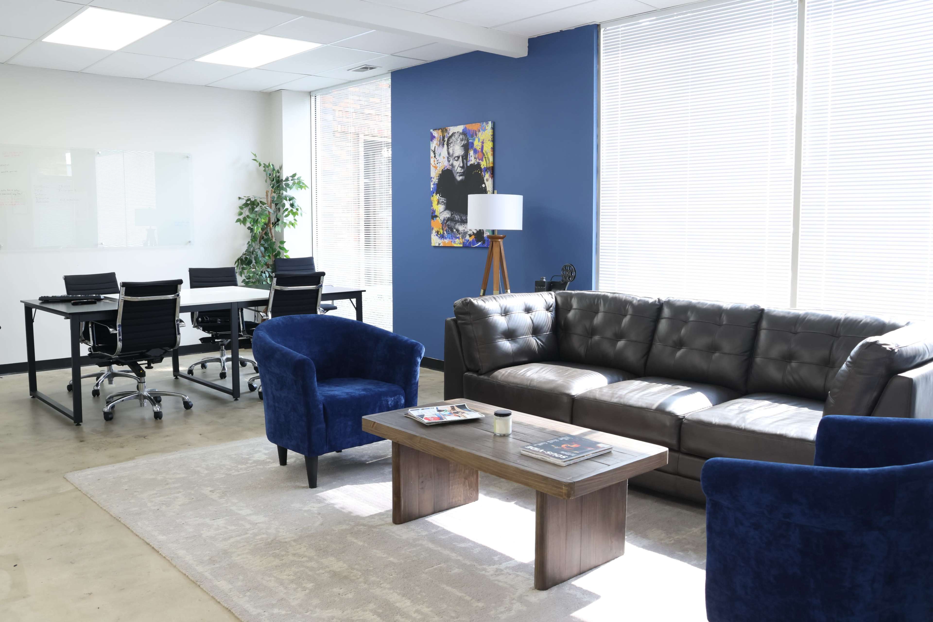 The modern office features a seating area with a brown leather sofa and two blue chairs, alongside a wooden coffee table, contrasting with a workspace equipped with a glass table and black chairs.