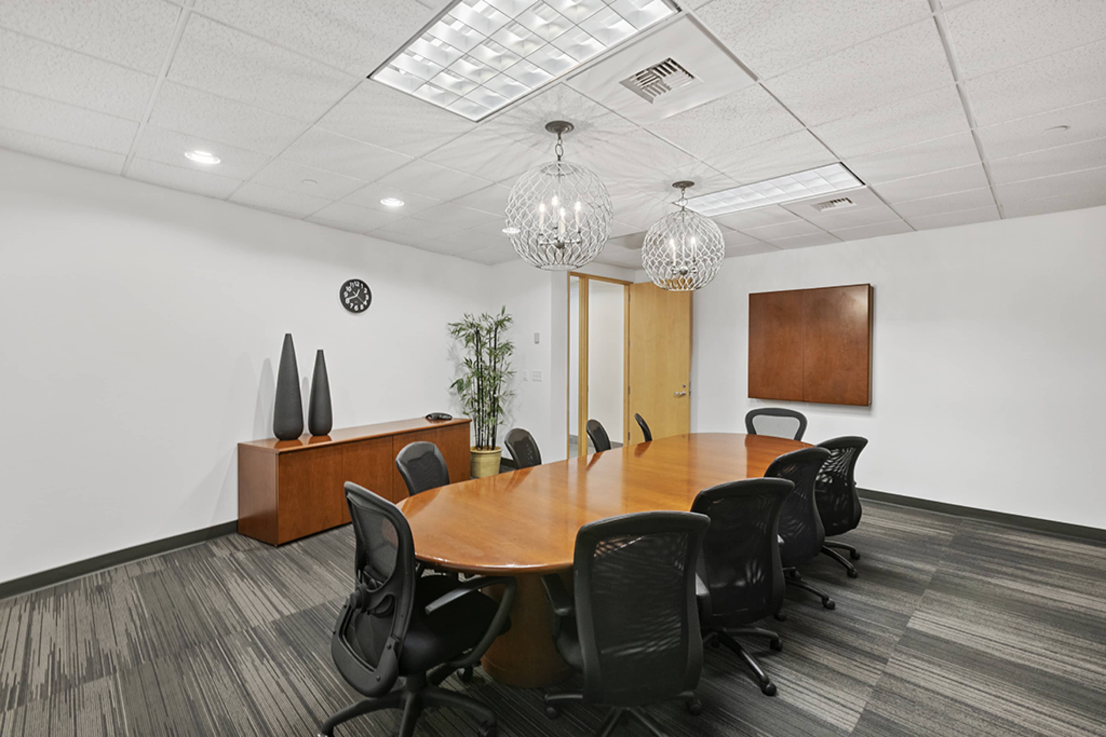 A modern conference room features a large wooden table surrounded by black chairs, with pendant lights and minimal decor.