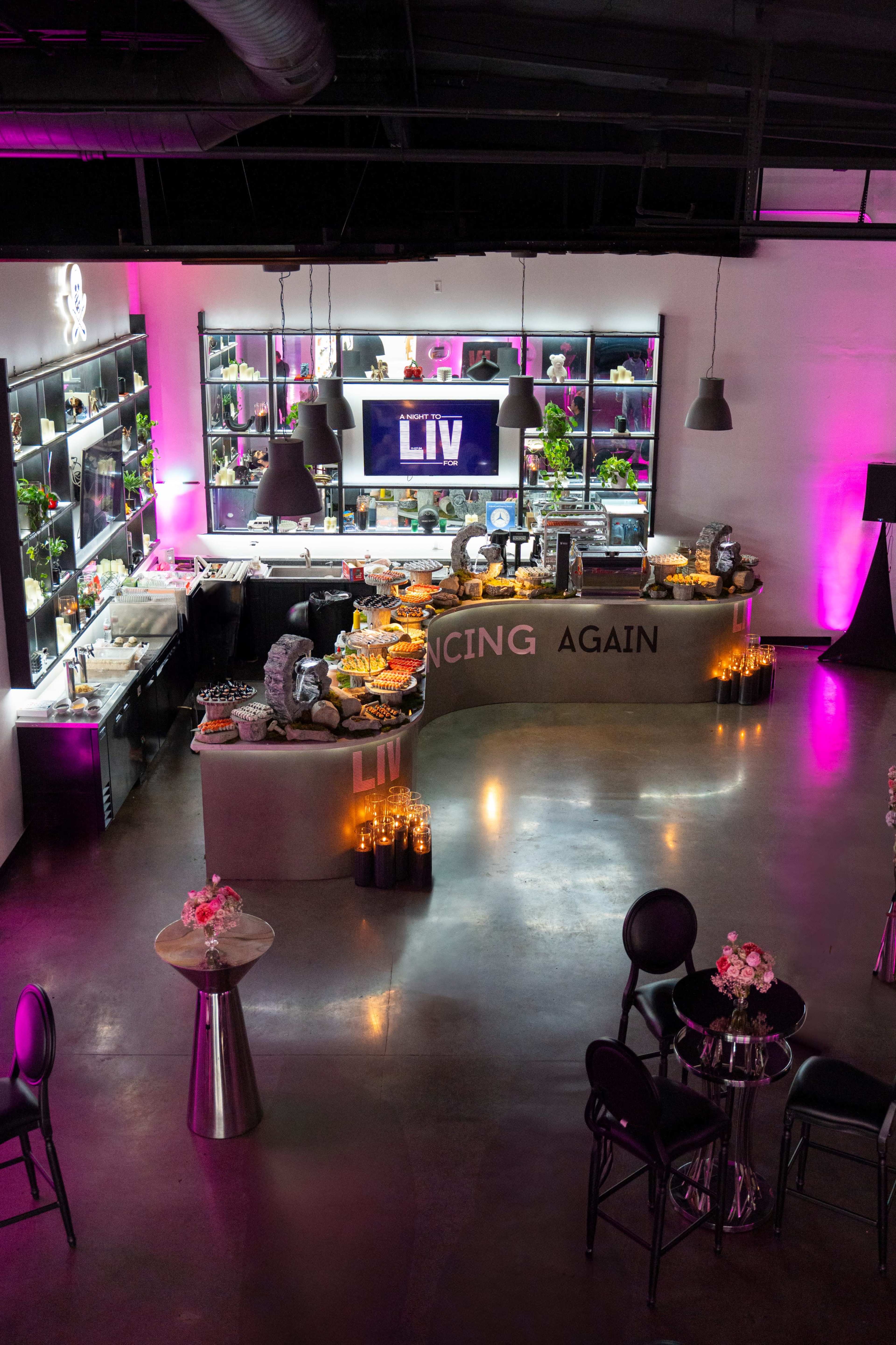 The image shows a modern event space with a curved bar display, buffet setup, and decorative lighting, featuring the word "LIV" prominently displayed.