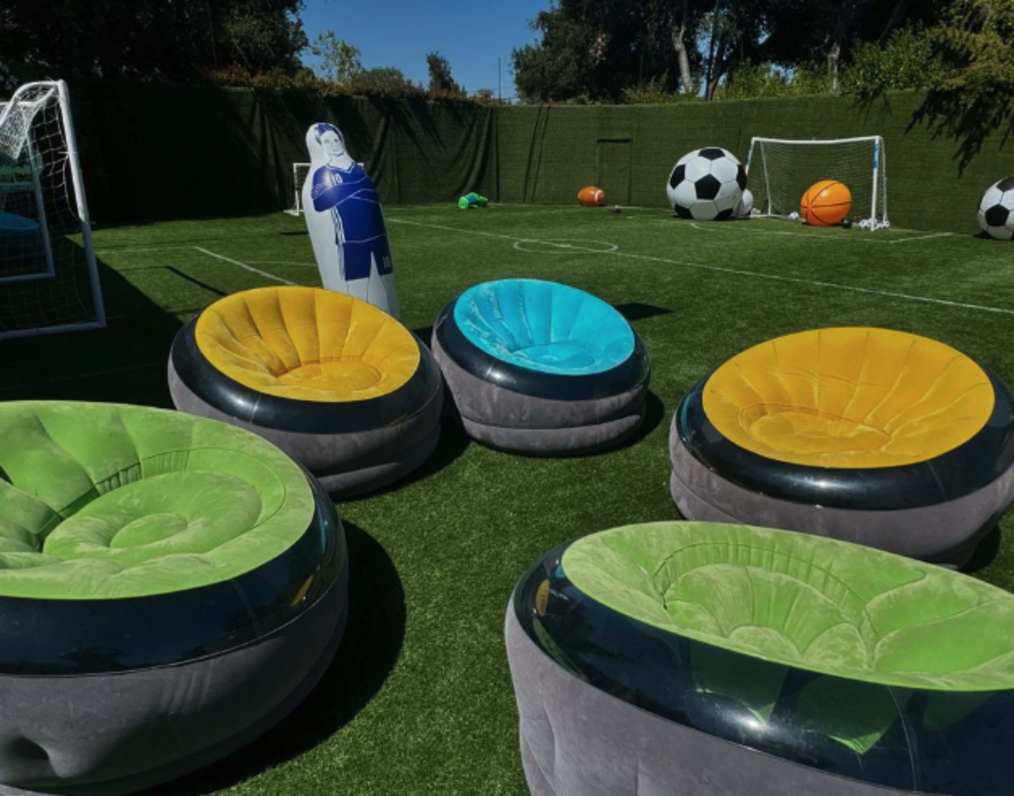 Five inflatable chairs in bright colors arranged on a grassy field with sports equipment, including soccer nets and large balls.