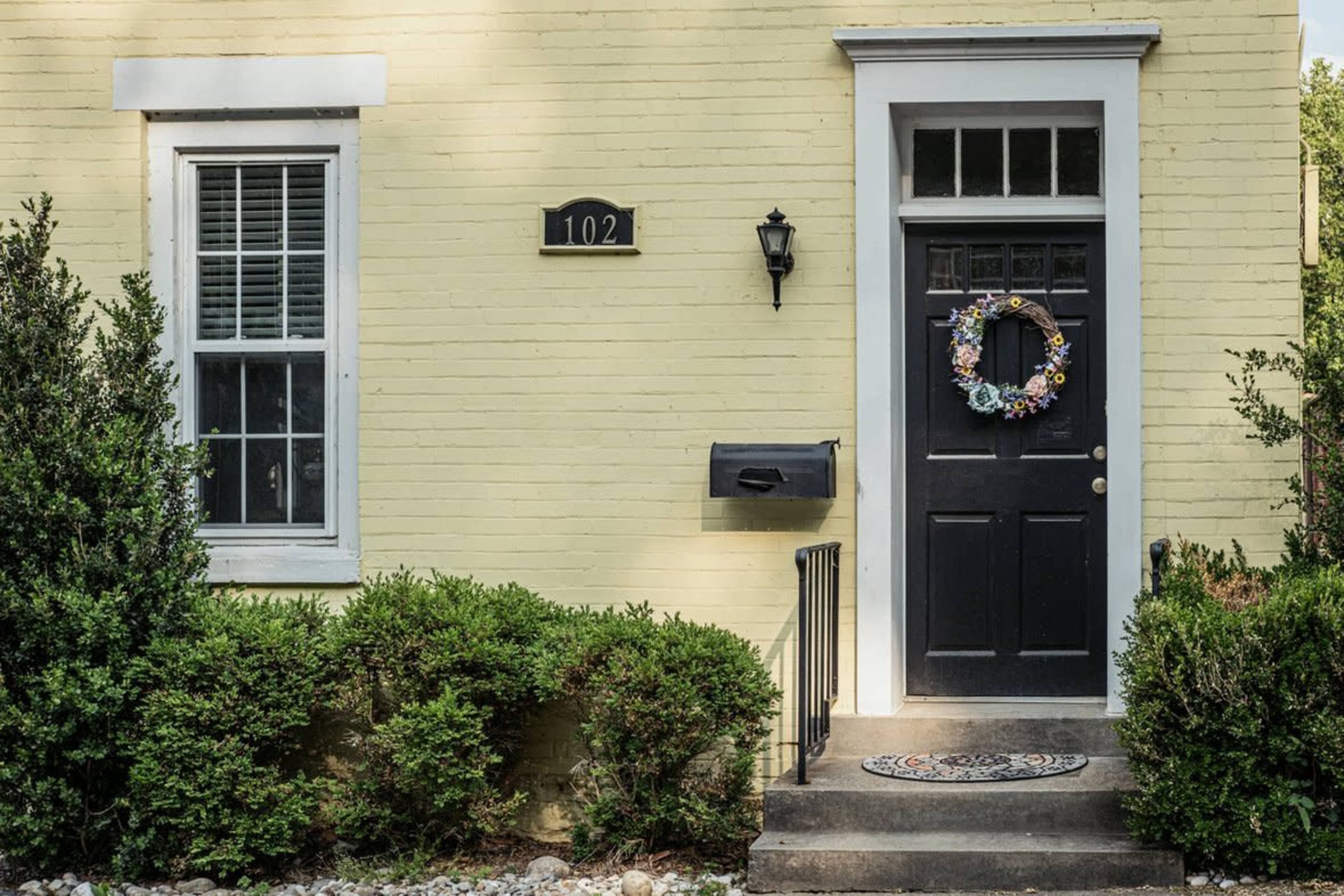 The image shows a yellow brick house with a black front door, a wreath, and a mailbox beside the entrance.