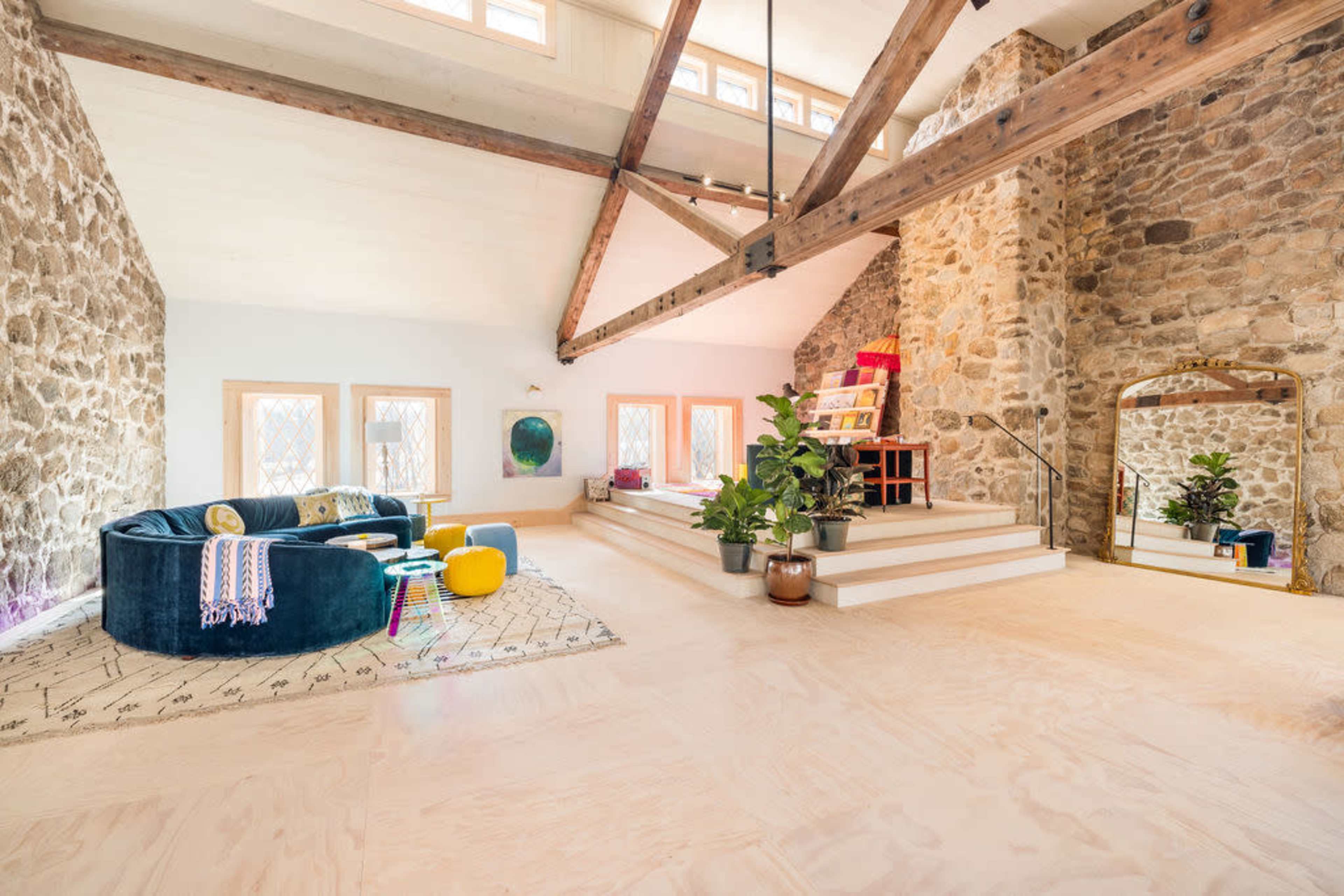 The interior features a spacious room with stone walls, a high ceiling supported by wooden beams, and a cozy seating area alongside decorative plants and stairs leading to a raised platform.