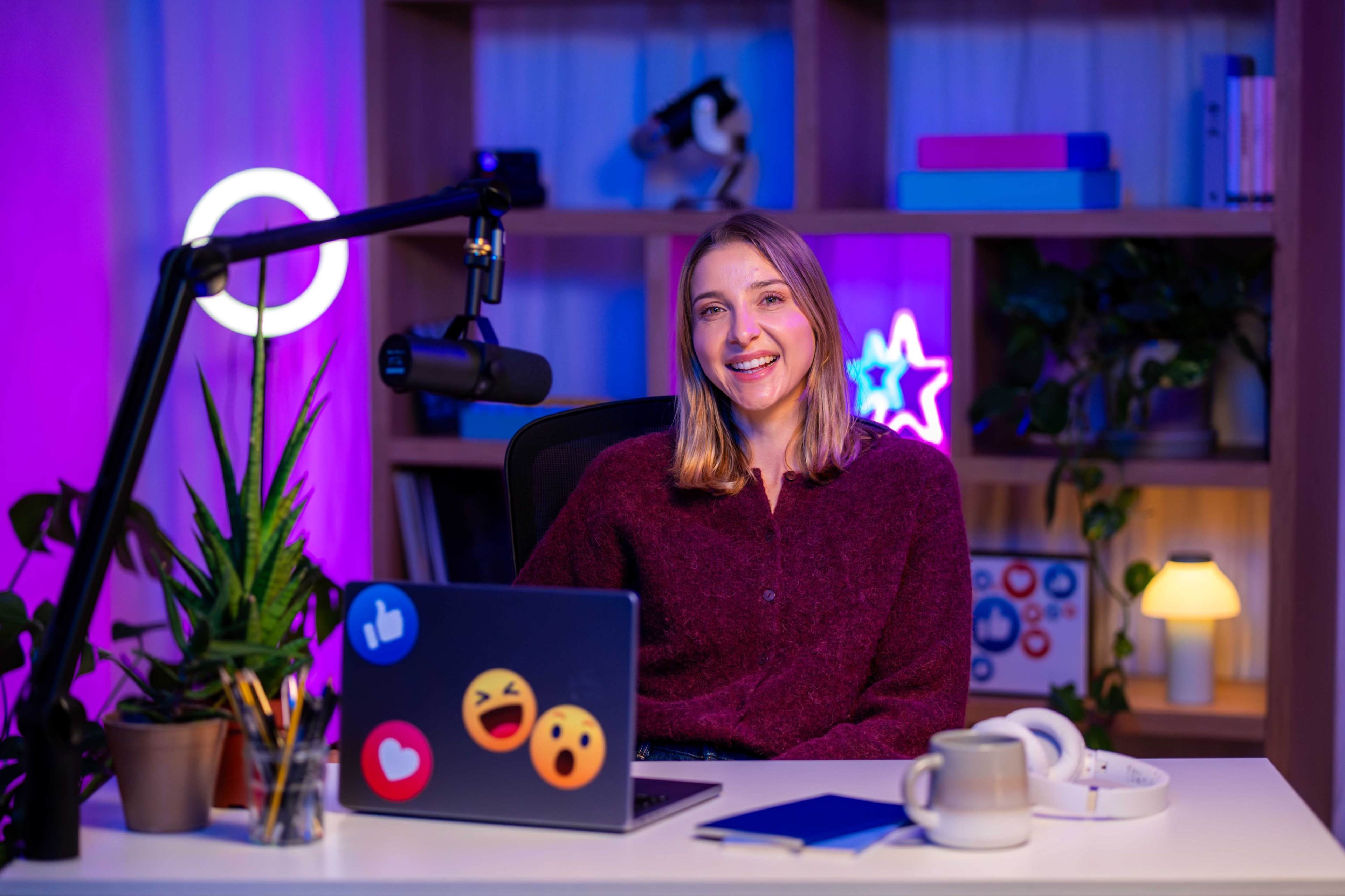 A young woman smiles while sitting at a desk with a laptop, plants, and colorful lighting in a modern workspace.