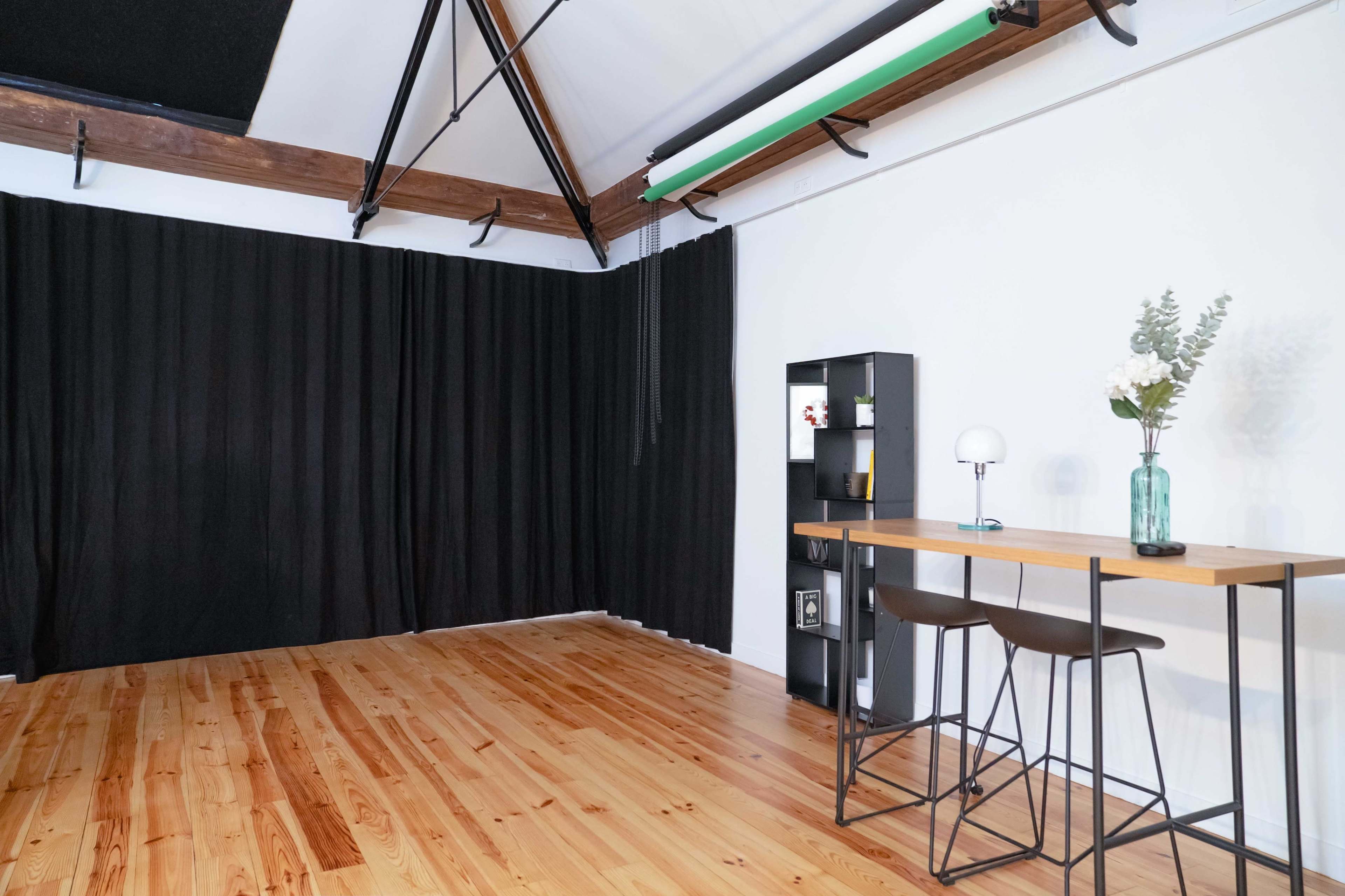 A minimalistic room features a wooden floor, a small table with two chairs, a black curtain, and a bookshelf with decorative items.