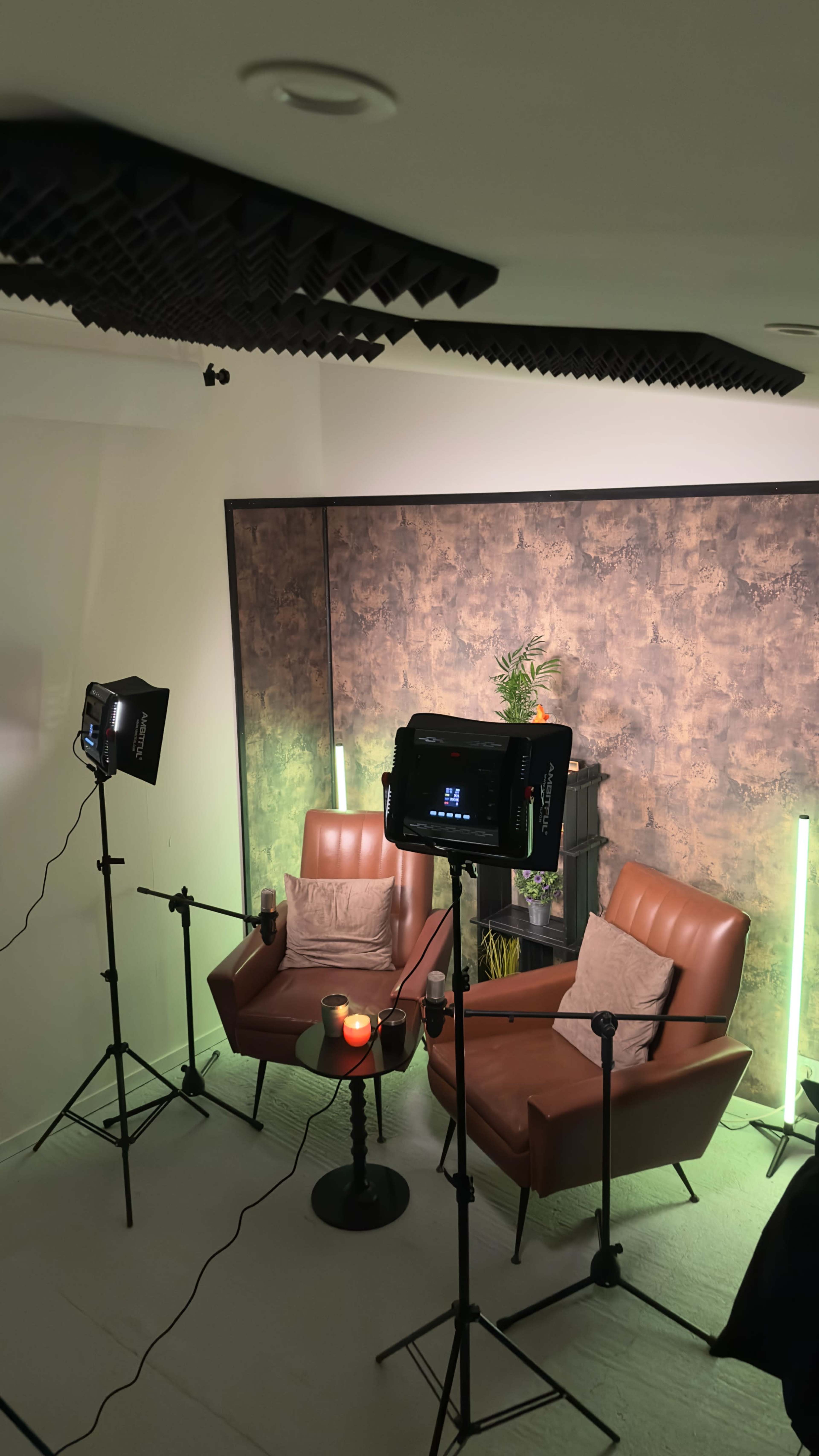 A small studio setup features two brown leather armchairs, a round table with a lit candle, and lighting equipment positioned on stands around the area.