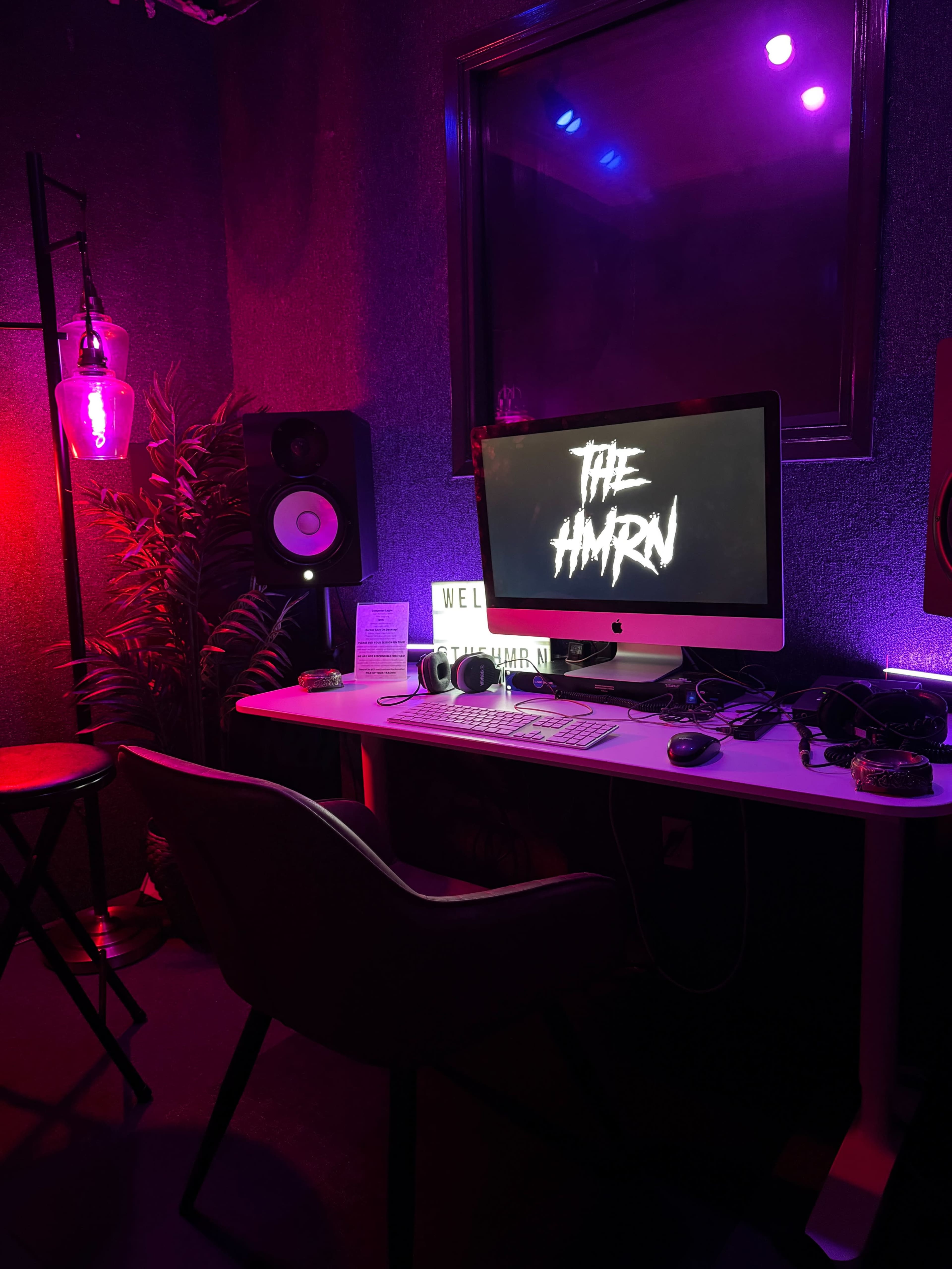 A modern computer desk is set up in a dimly lit room with colorful lighting, featuring a large monitor displaying "THE HAMRN," surrounded by audio equipment and decorative plants.