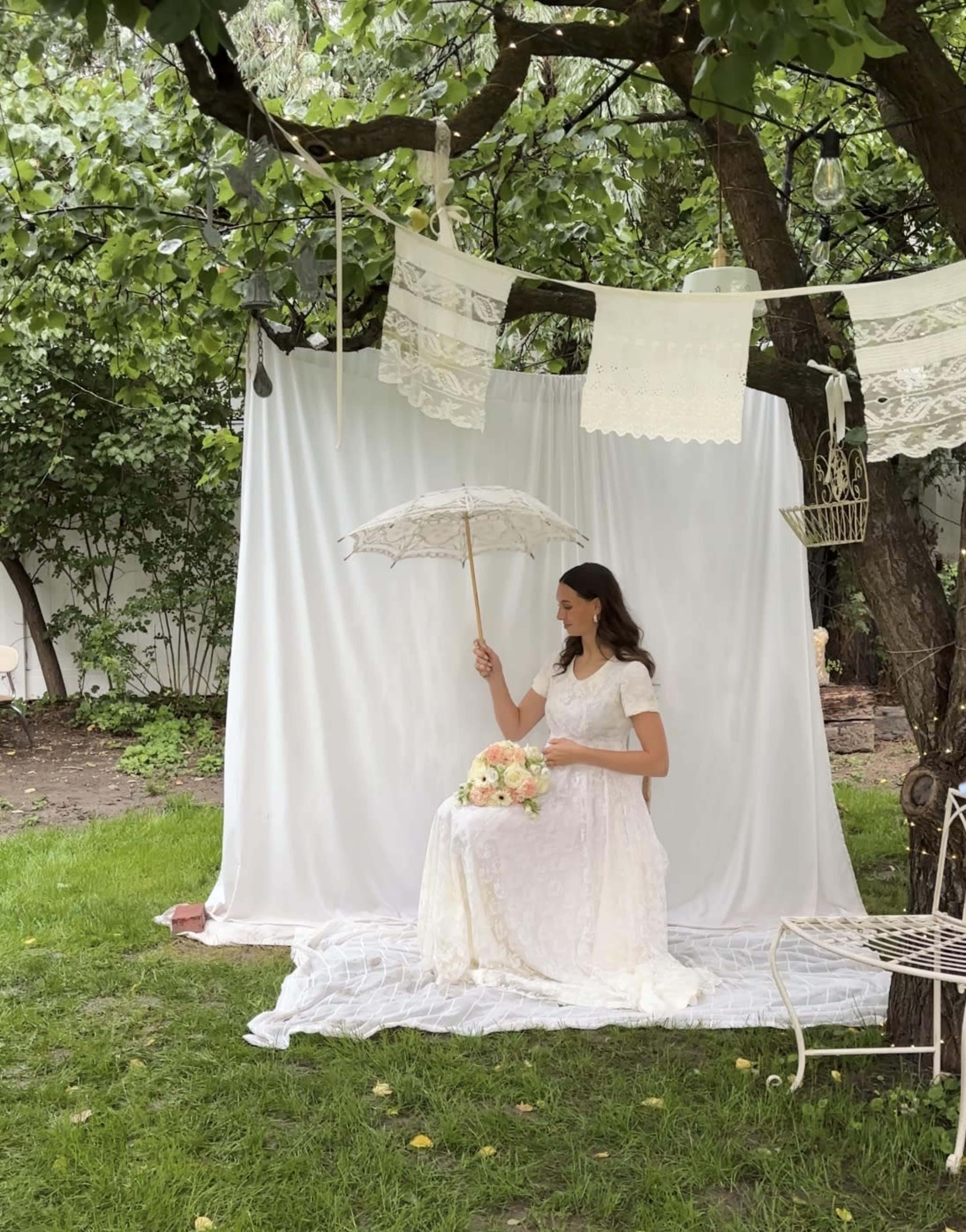 A woman in a white dress sits on a blanket in a garden, holding a parasol, with white fabric draped as a backdrop and decorative items hanging from nearby branches.