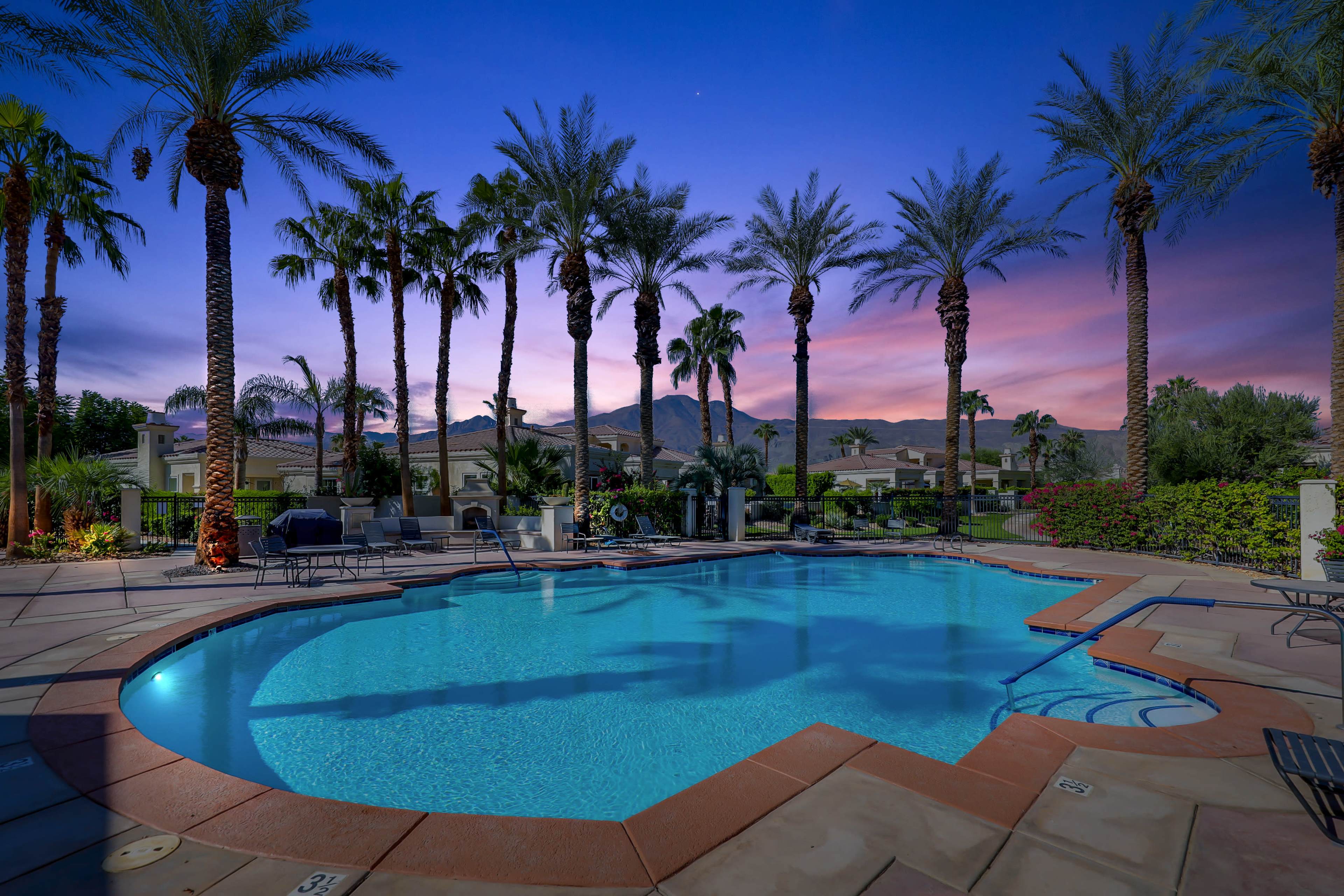 The image shows a swimming pool surrounded by palm trees and landscaped homes under a twilight sky.