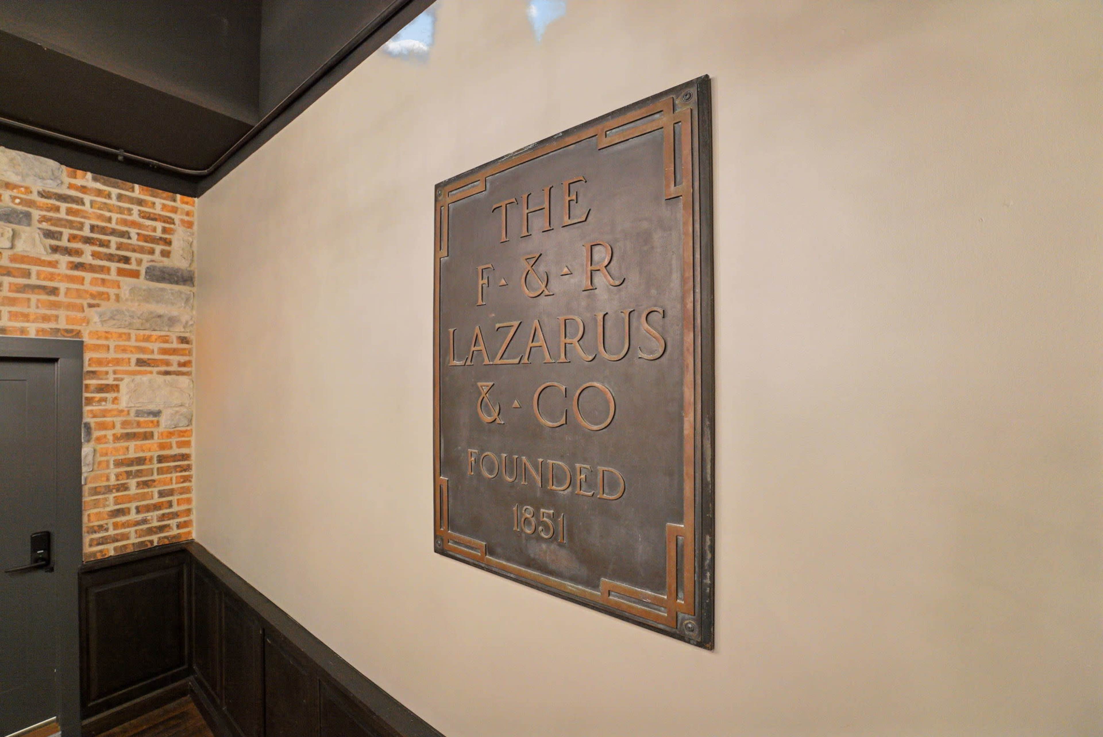 The image shows a wall-mounted plaque that reads "THE F & R LAZARUS & CO FOUNDED 1851" in an industrial-style setting with a textured wall.