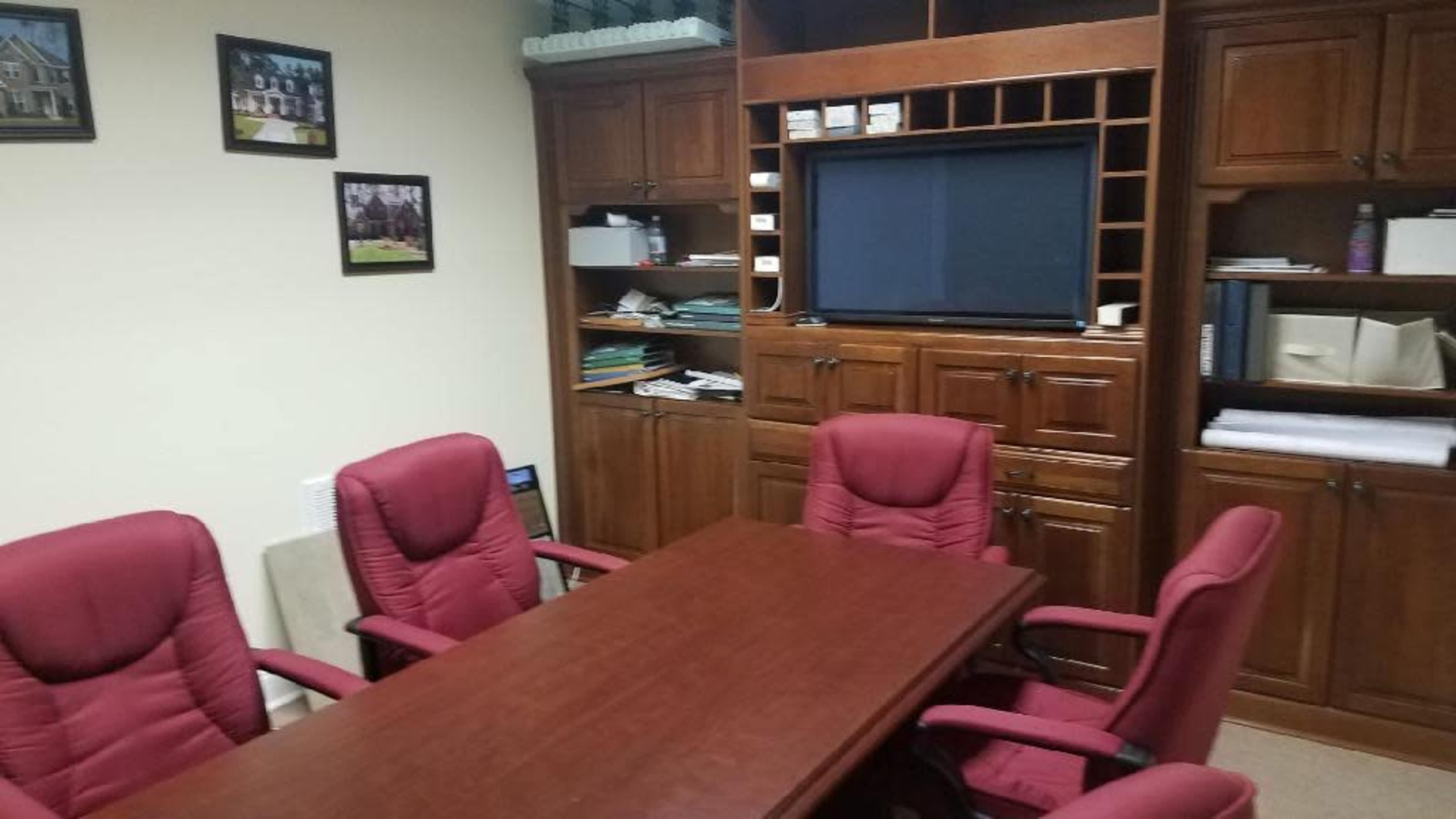 A conference room with a rectangular wooden table surrounded by five red upholstered chairs, set against a backdrop of wooden cabinets and a wall-mounted screen.