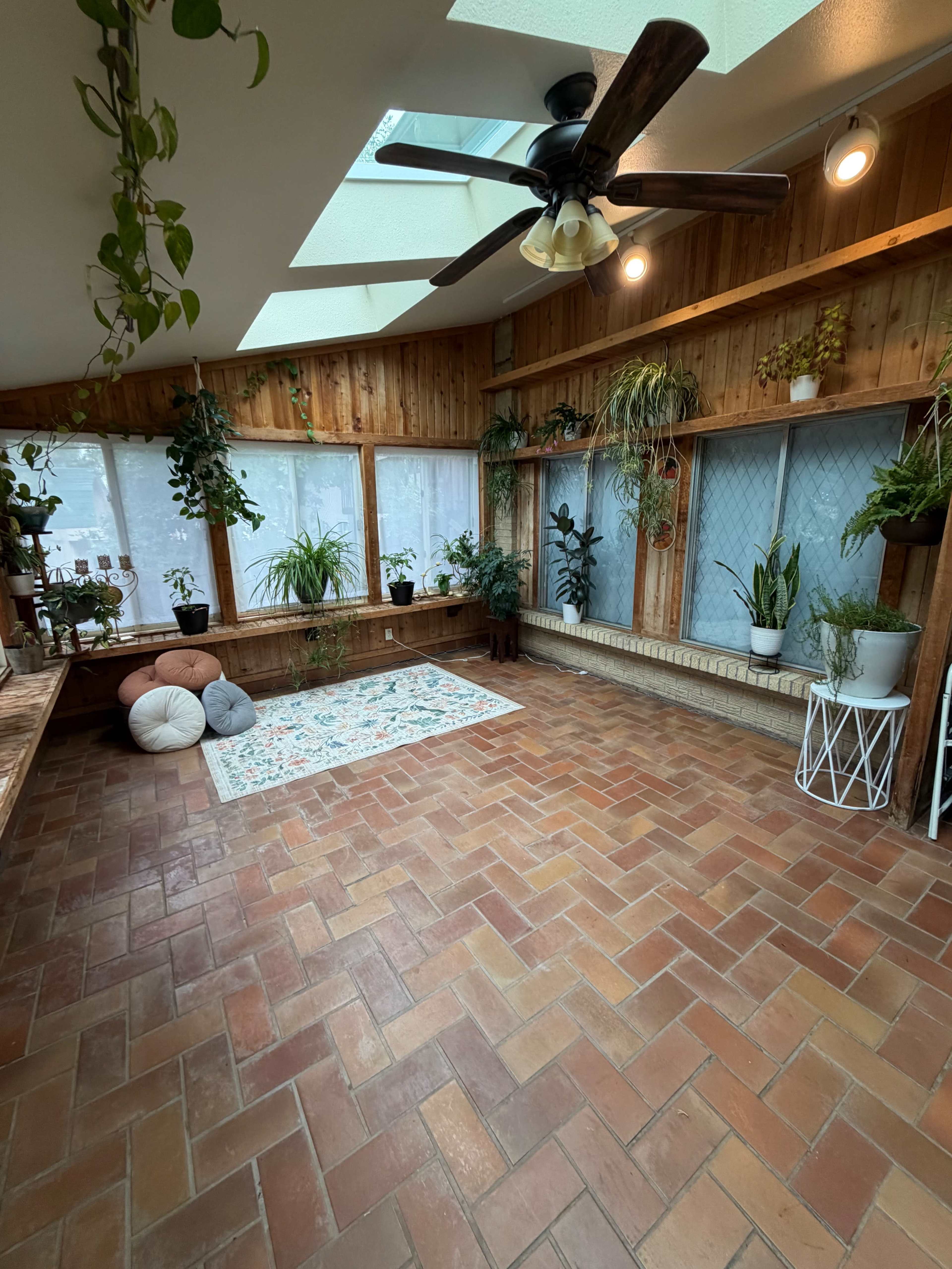 The image shows a sunroom with terracotta tile flooring, large windows lined with various potted plants, and a white rug in the center.