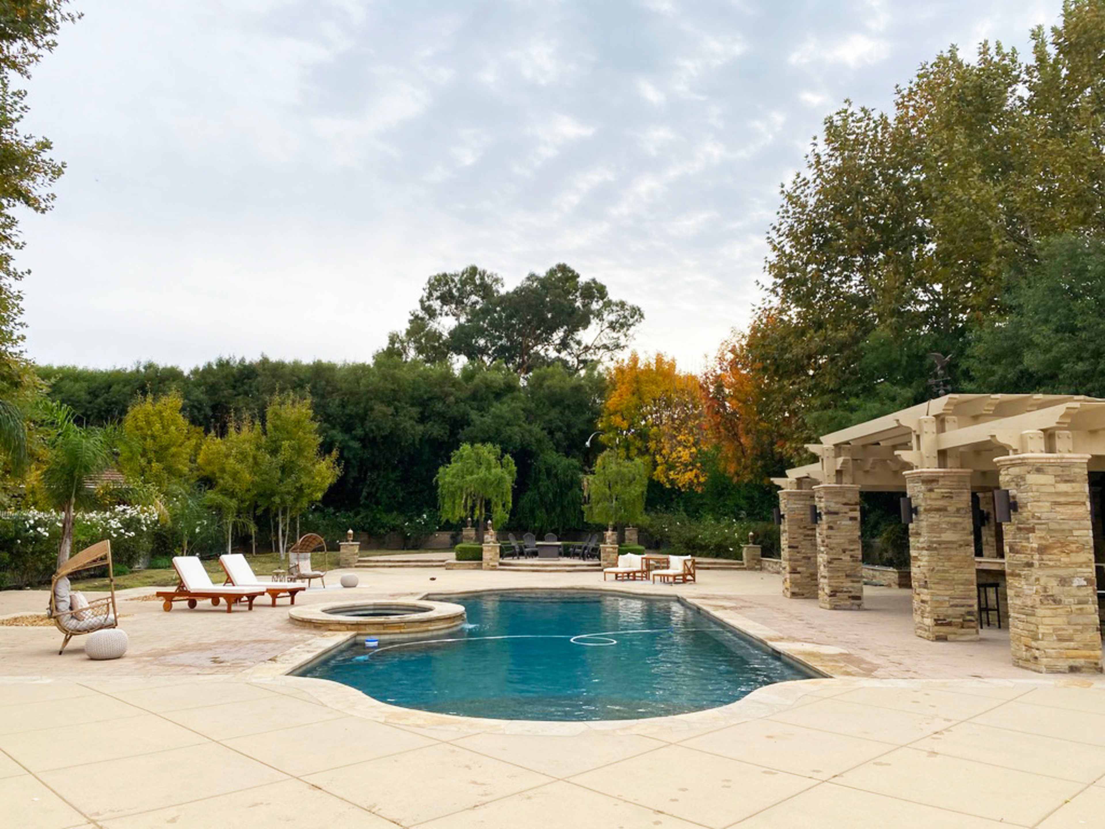 A backyard pool area surrounded by trees, lounge chairs, and a covered seating space.