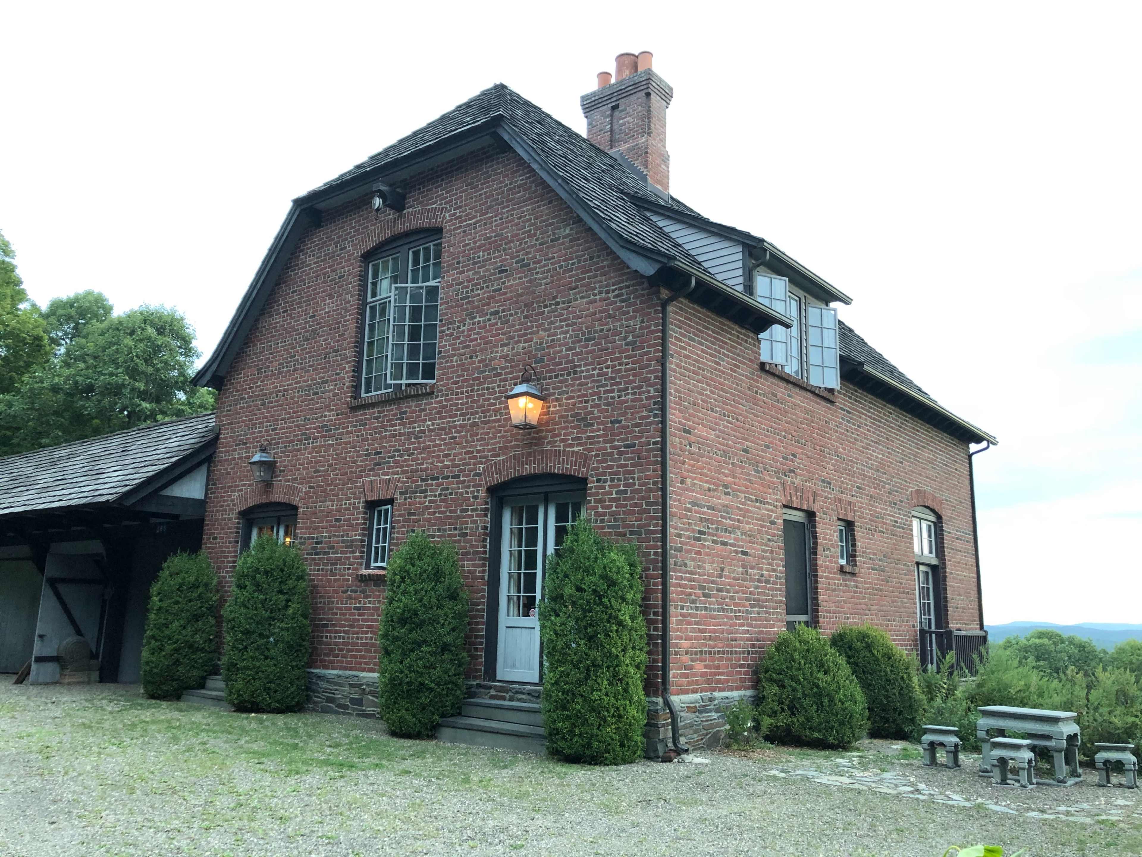 A two-story brick house with a chimney and several shrubs in front is situated on a gravel path.