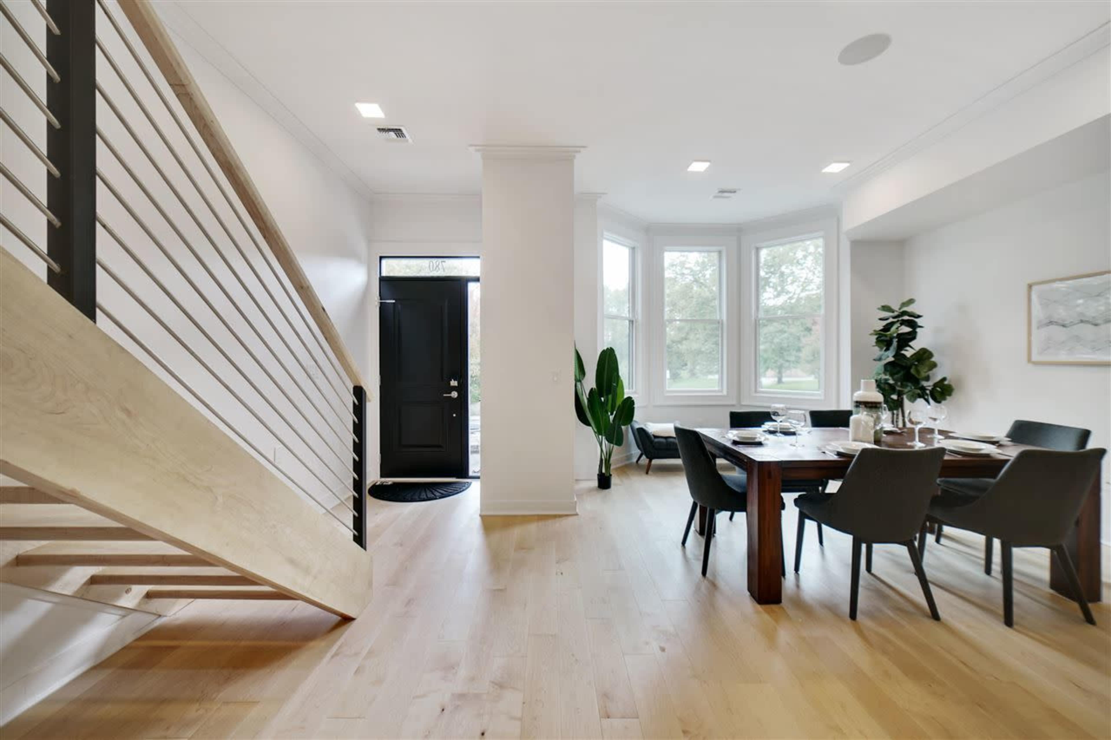 A modern entryway and dining area with a wooden staircase, a round dining table, and large windows allowing natural light to fill the space.