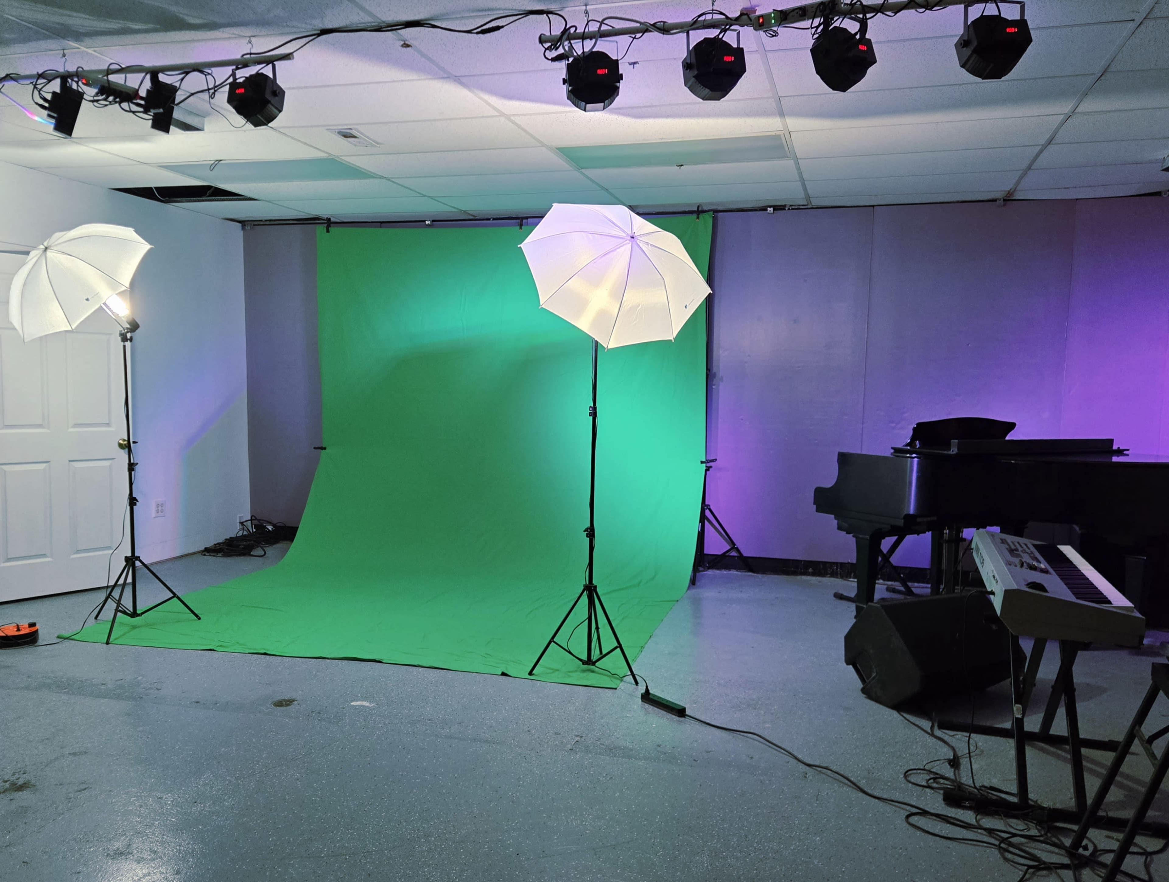 The image shows a photography studio with a green backdrop, two white umbrella lights, and a piano alongside a keyboard in the corner.
