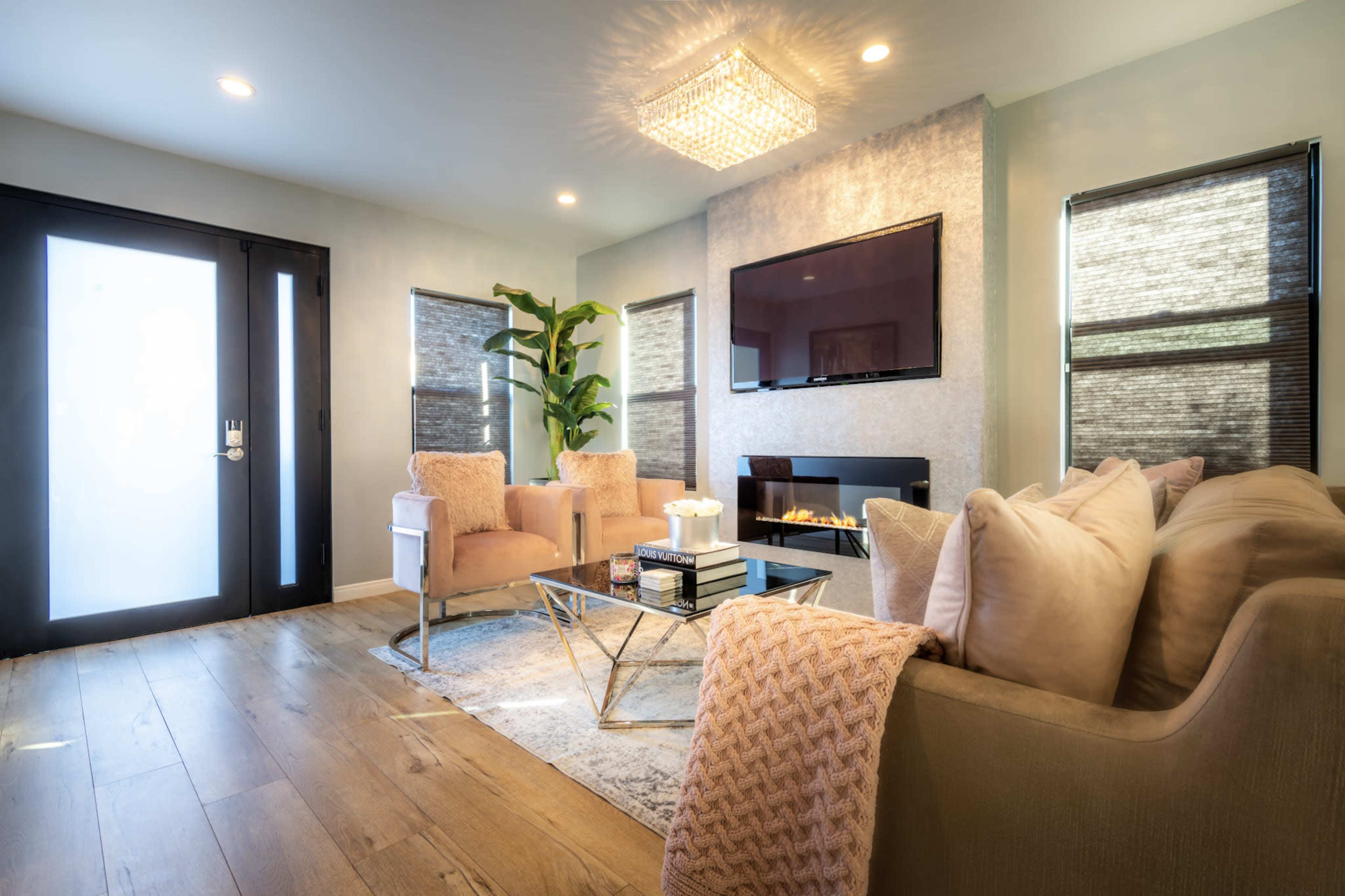 The image shows a modern living room featuring two armchairs, a coffee table, a television mounted on the wall, and large windows with natural light.