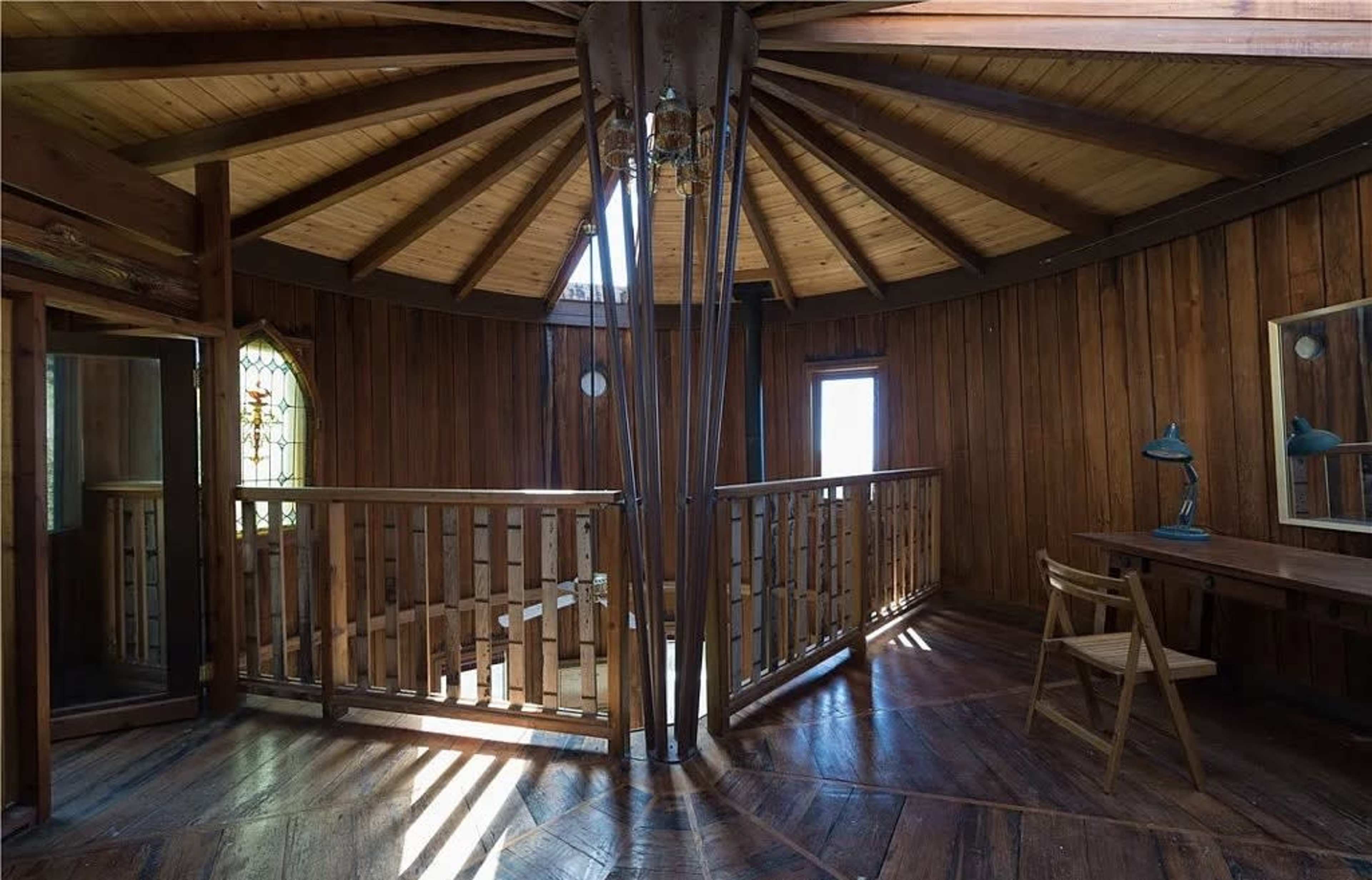 The interior of a circular wooden room features a staircase, large windows, and a desk with a lamp.