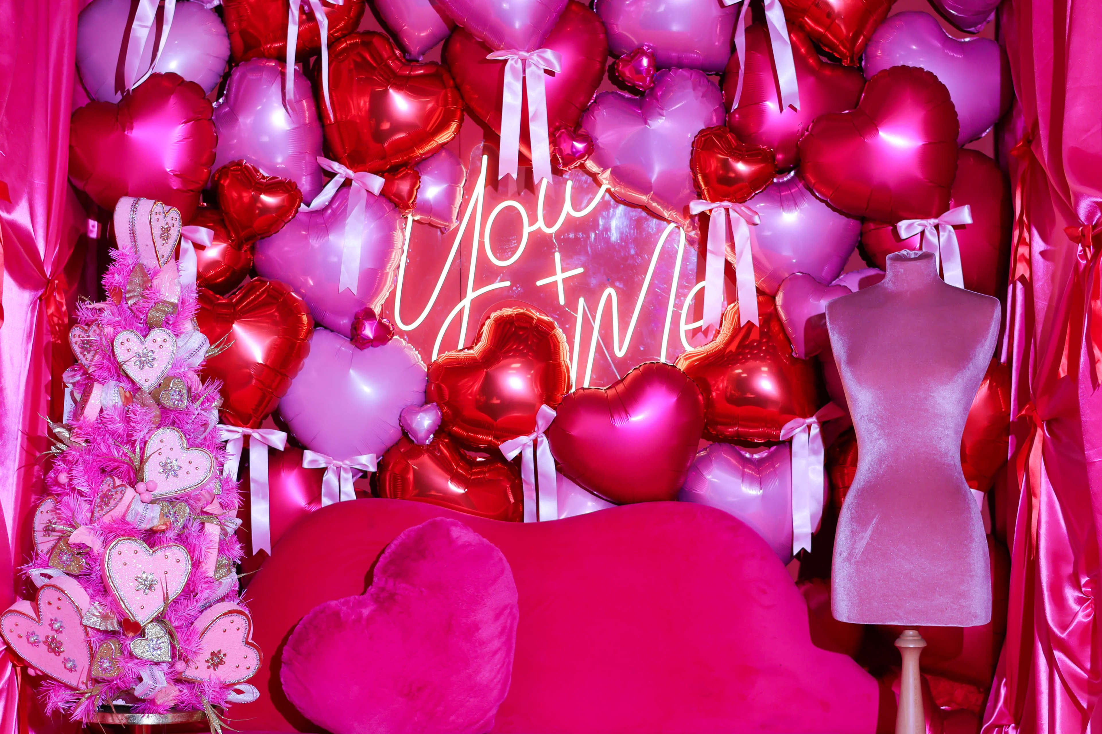 A bright pink backdrop displays a heart-themed arrangement of balloons and a neon sign reading "You + Me," alongside a decorative Christmas tree and a pink couch.