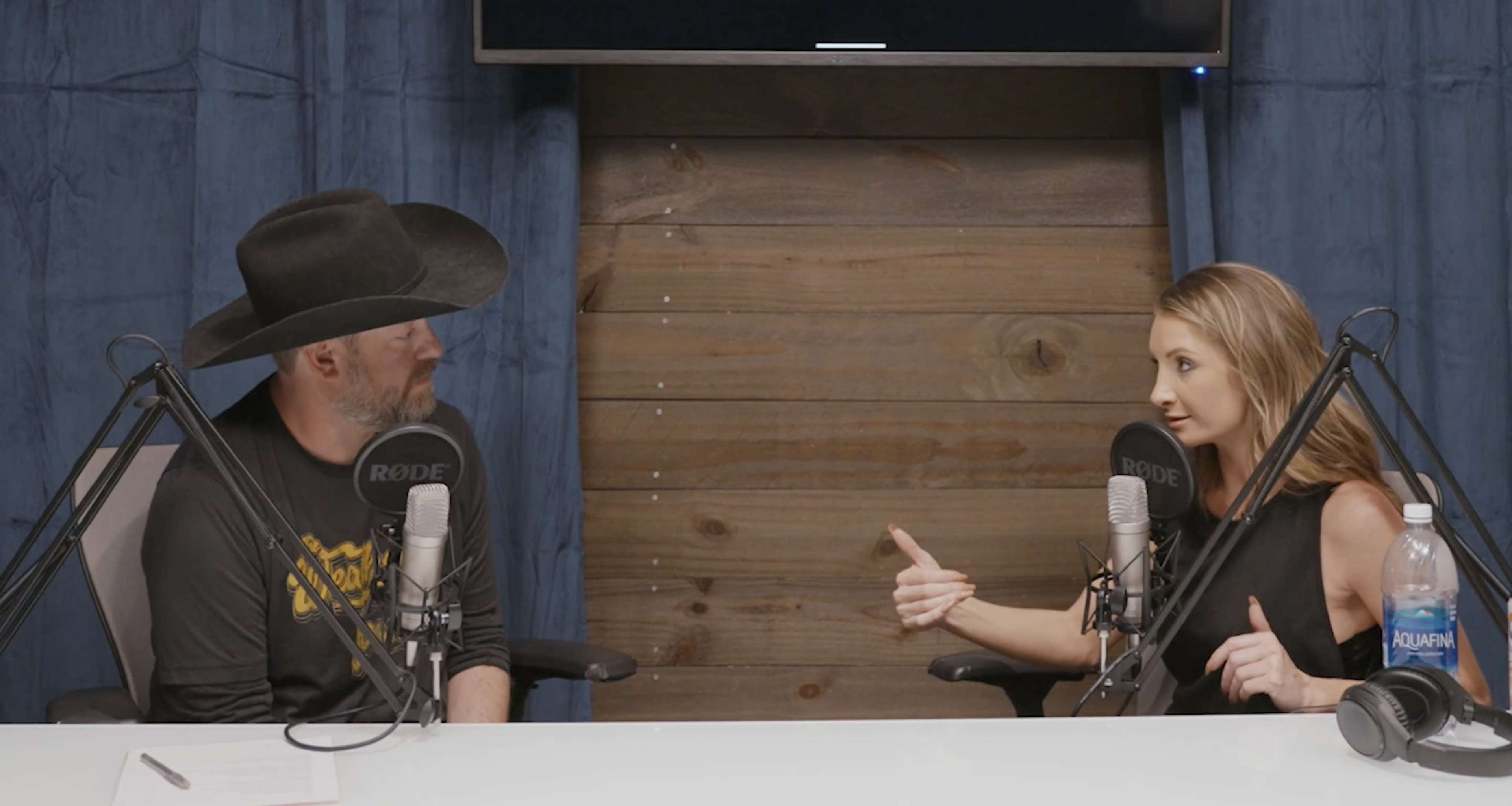 Two people are seated at a table with microphones in front of them, engaged in conversation against a backdrop of wooden panels and a digital screen.