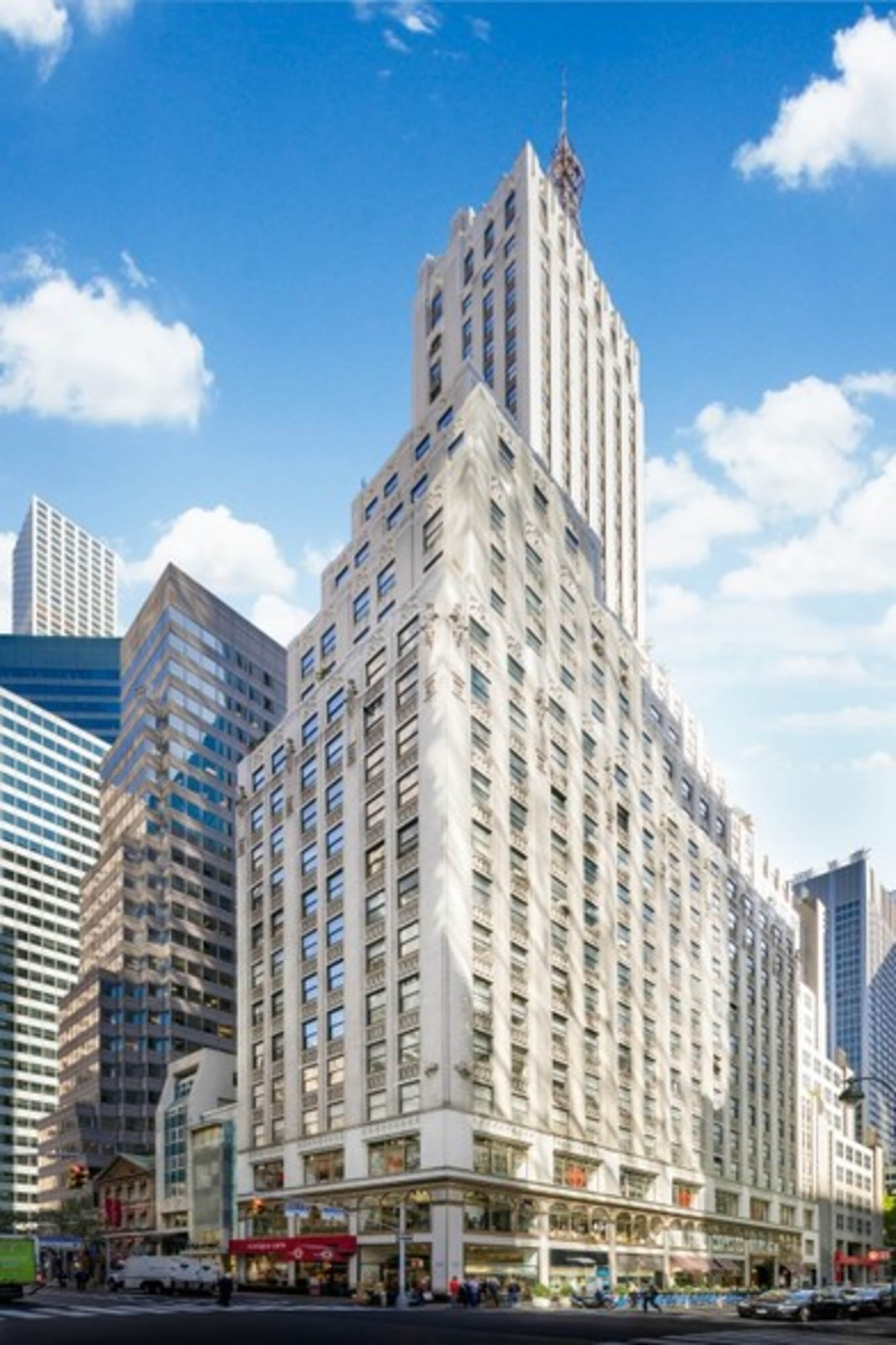 A tall, art deco building with a sharp, angled facade surrounded by modern skyscrapers under a blue sky.
