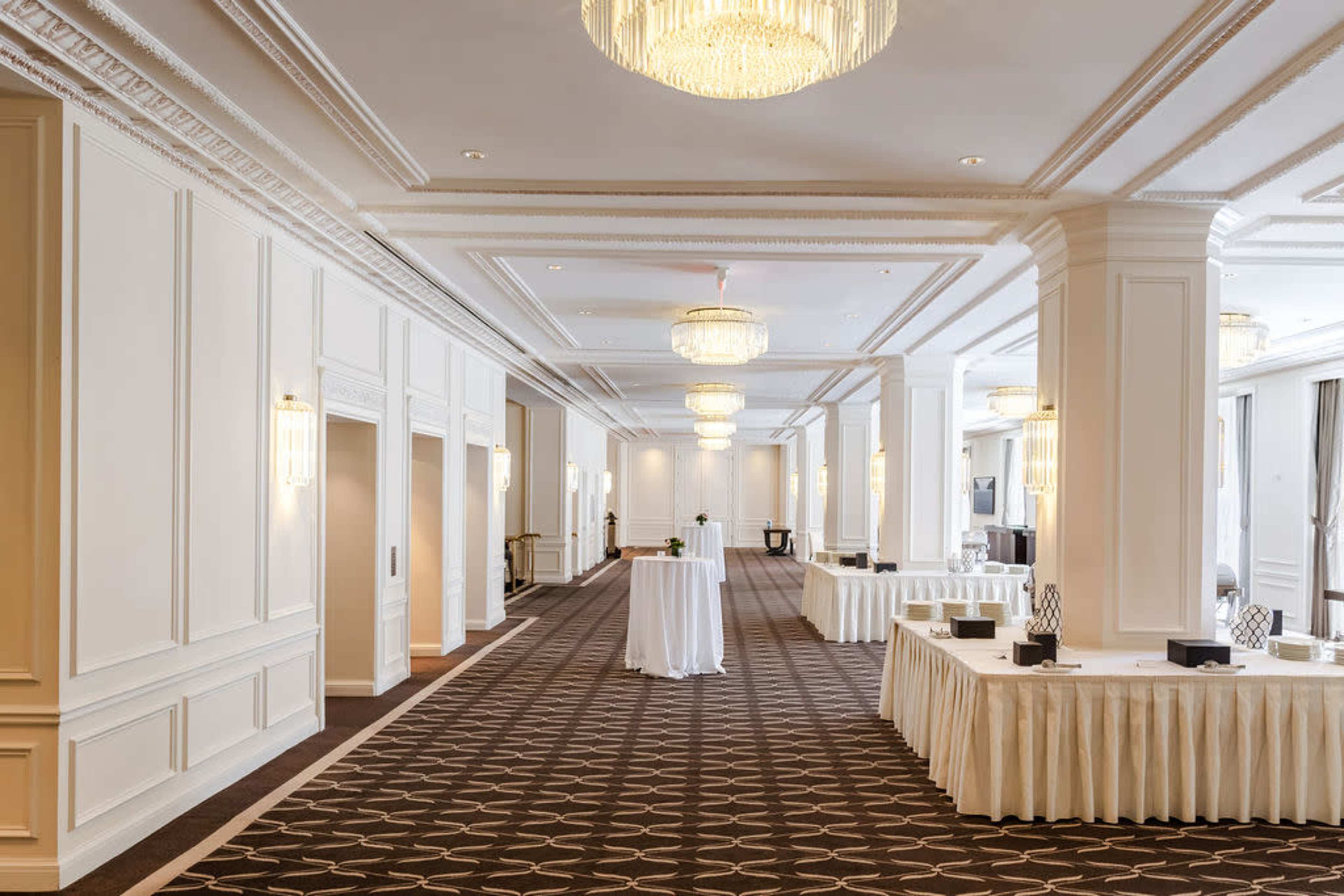 A spacious, elegantly decorated hallway features chandeliers, columns, and several tables arranged along the sides.