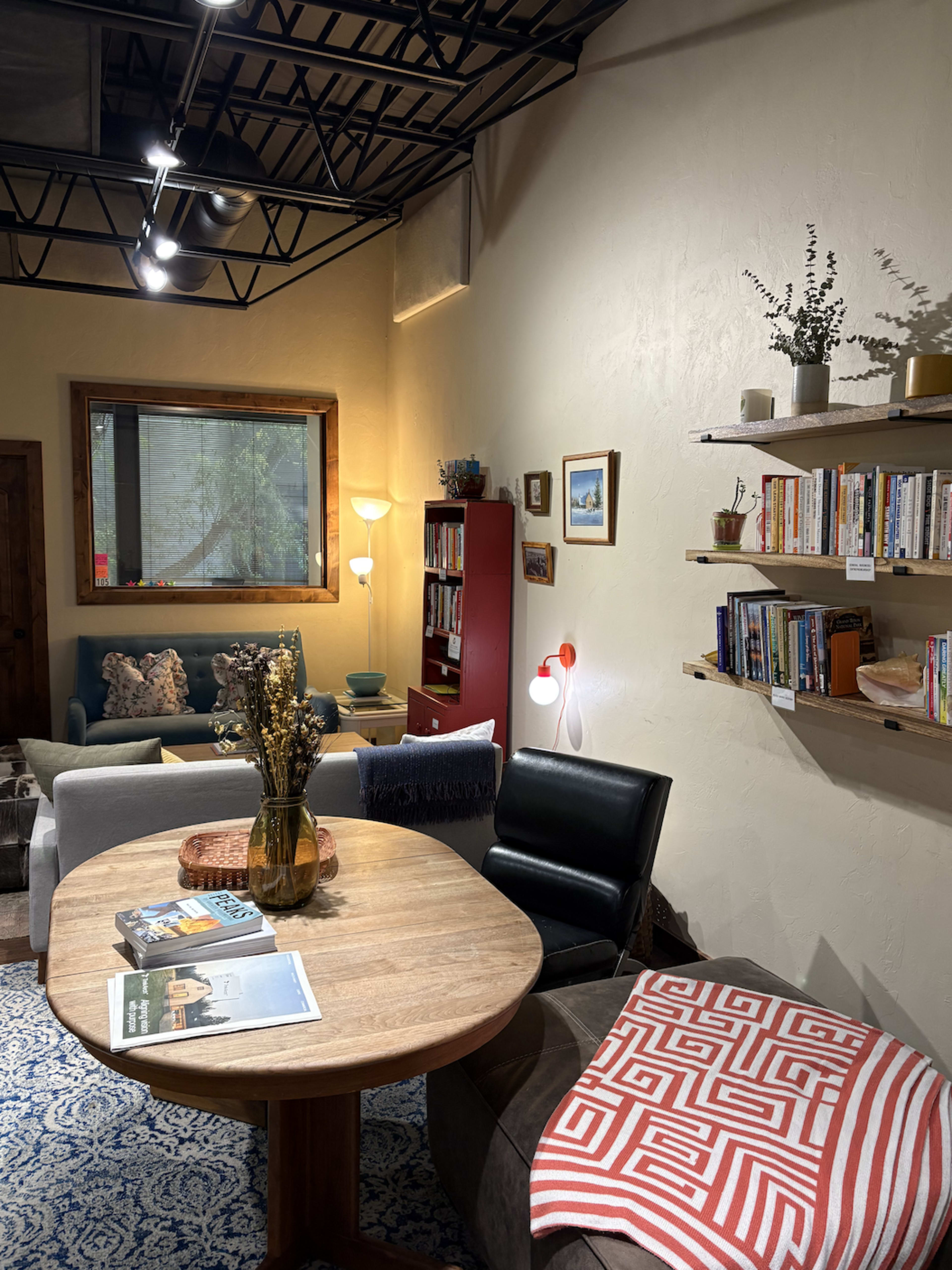 The image shows a cozy living space featuring a round wooden table, a black leather chair, a sofa with decorative pillows, and books arranged on shelves along the wall.