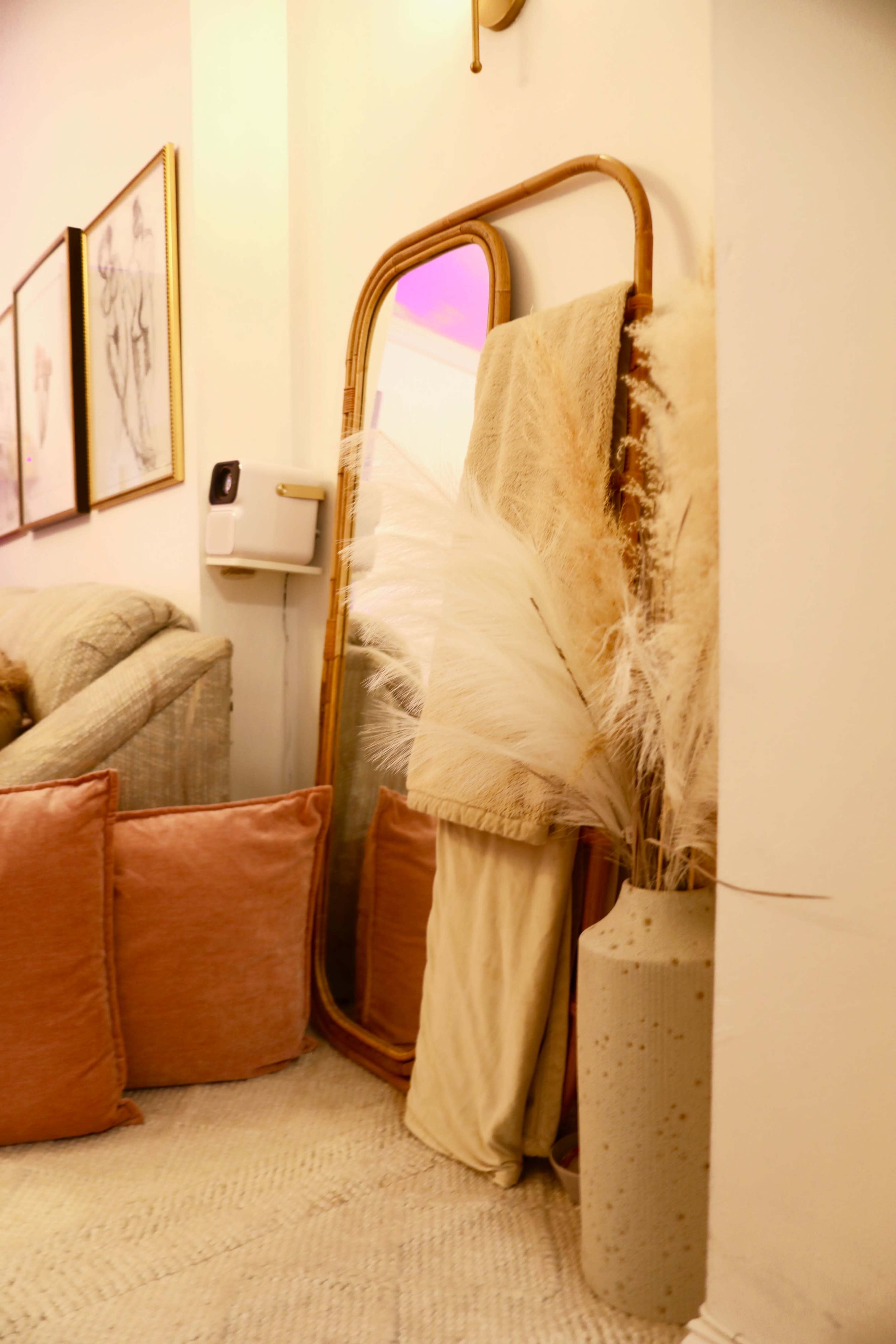 A large mirror is positioned against a wall next to a vase filled with pampas grass, with soft cushions arranged on a nearby sofa.