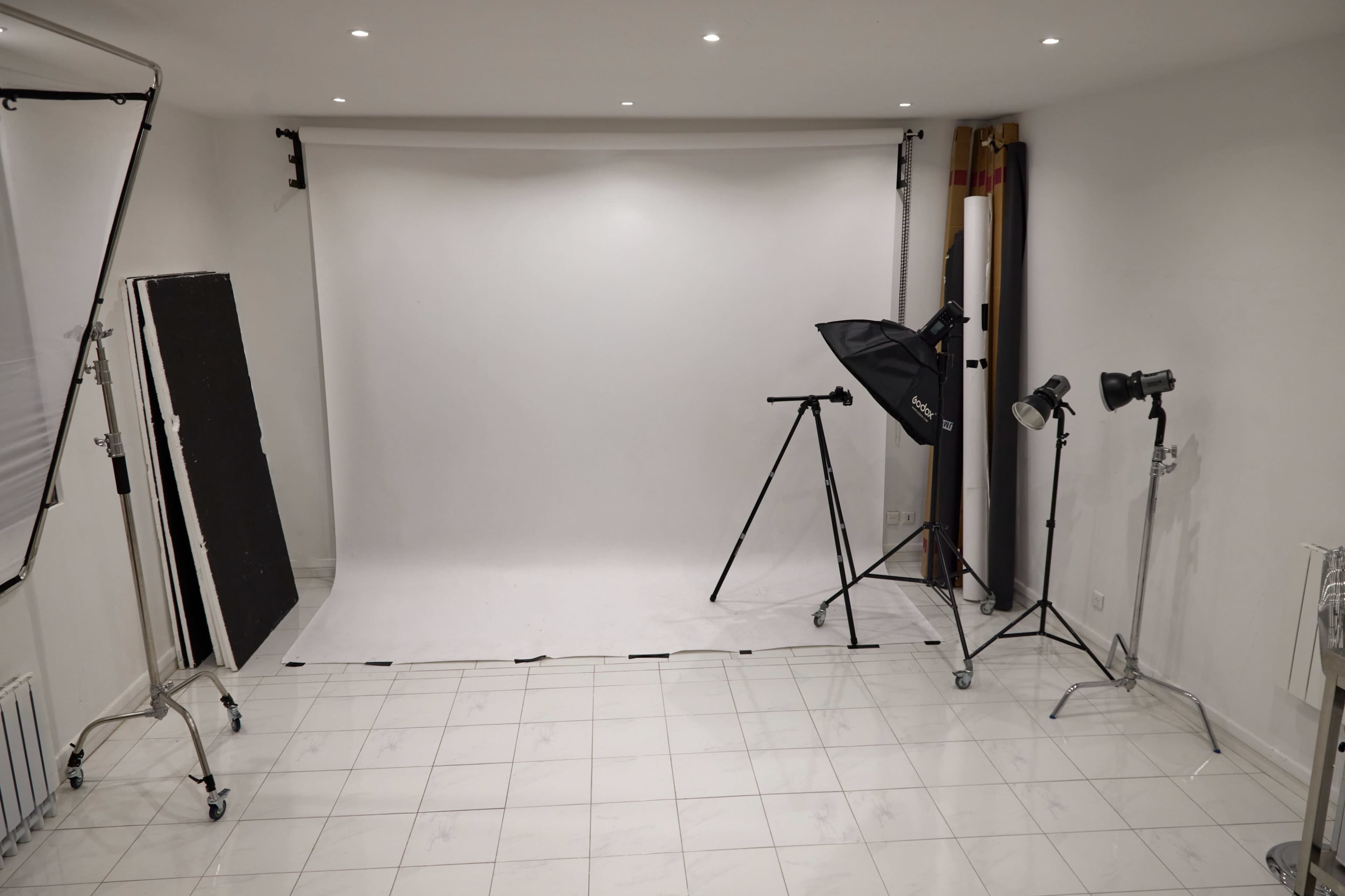 The image shows a photography studio with a white backdrop, softbox lights, and a tripod setup on a tiled floor.
