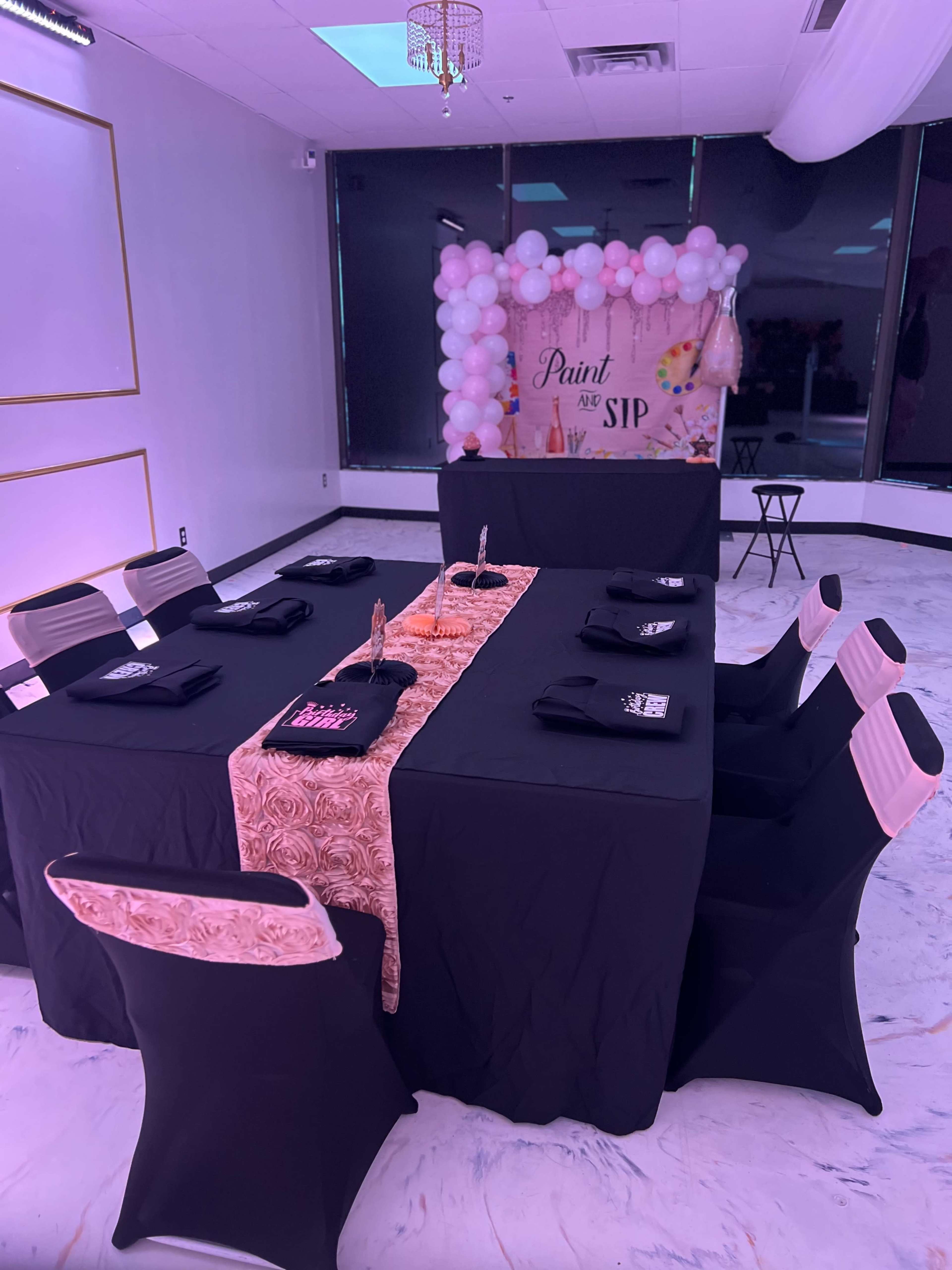 A black and gold decorated table is set in a brightly lit room with a backdrop of balloons and a sign for a "Paint and Sip" event.