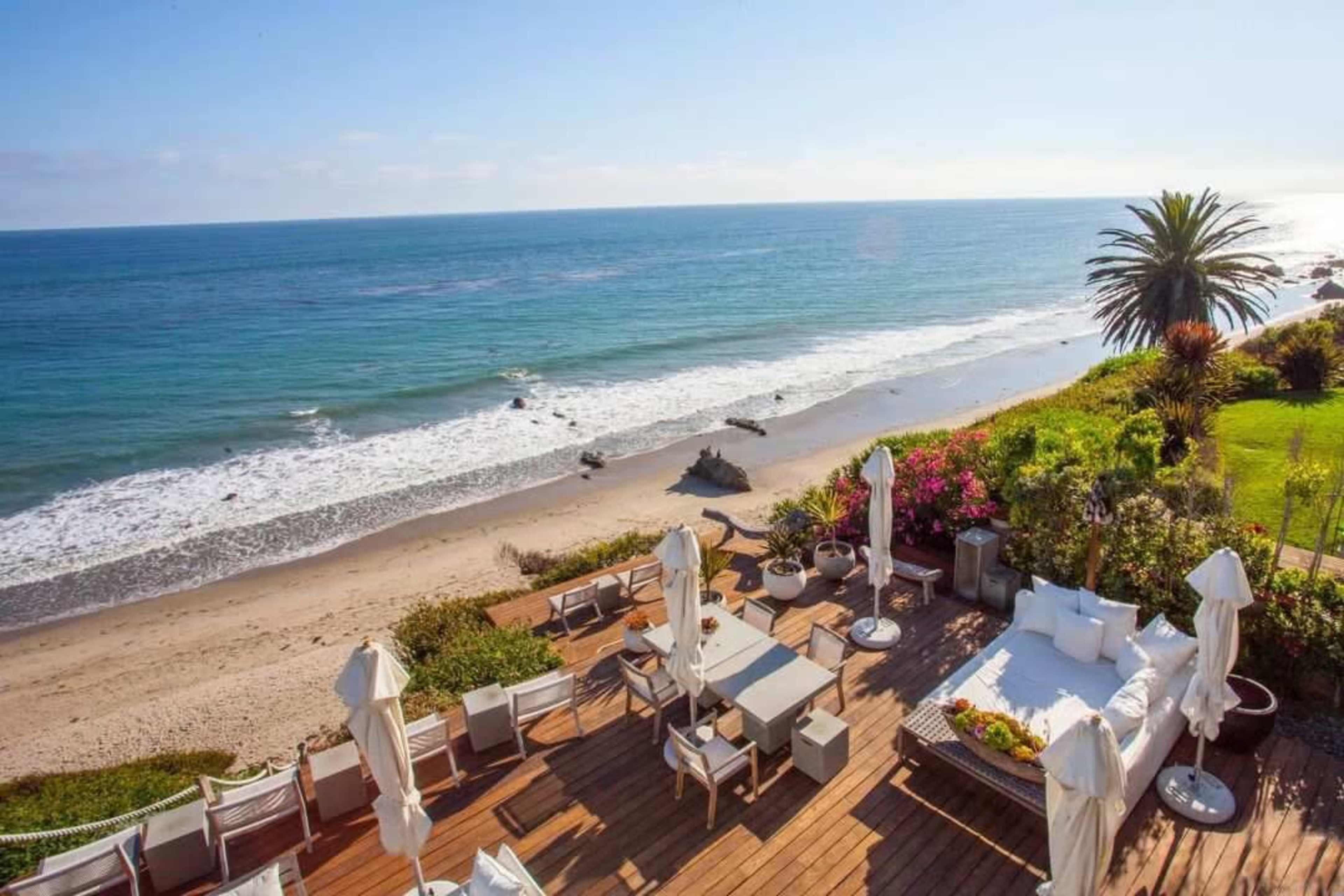 A deck with outdoor furniture overlooks a sandy beach and ocean against a clear blue sky.