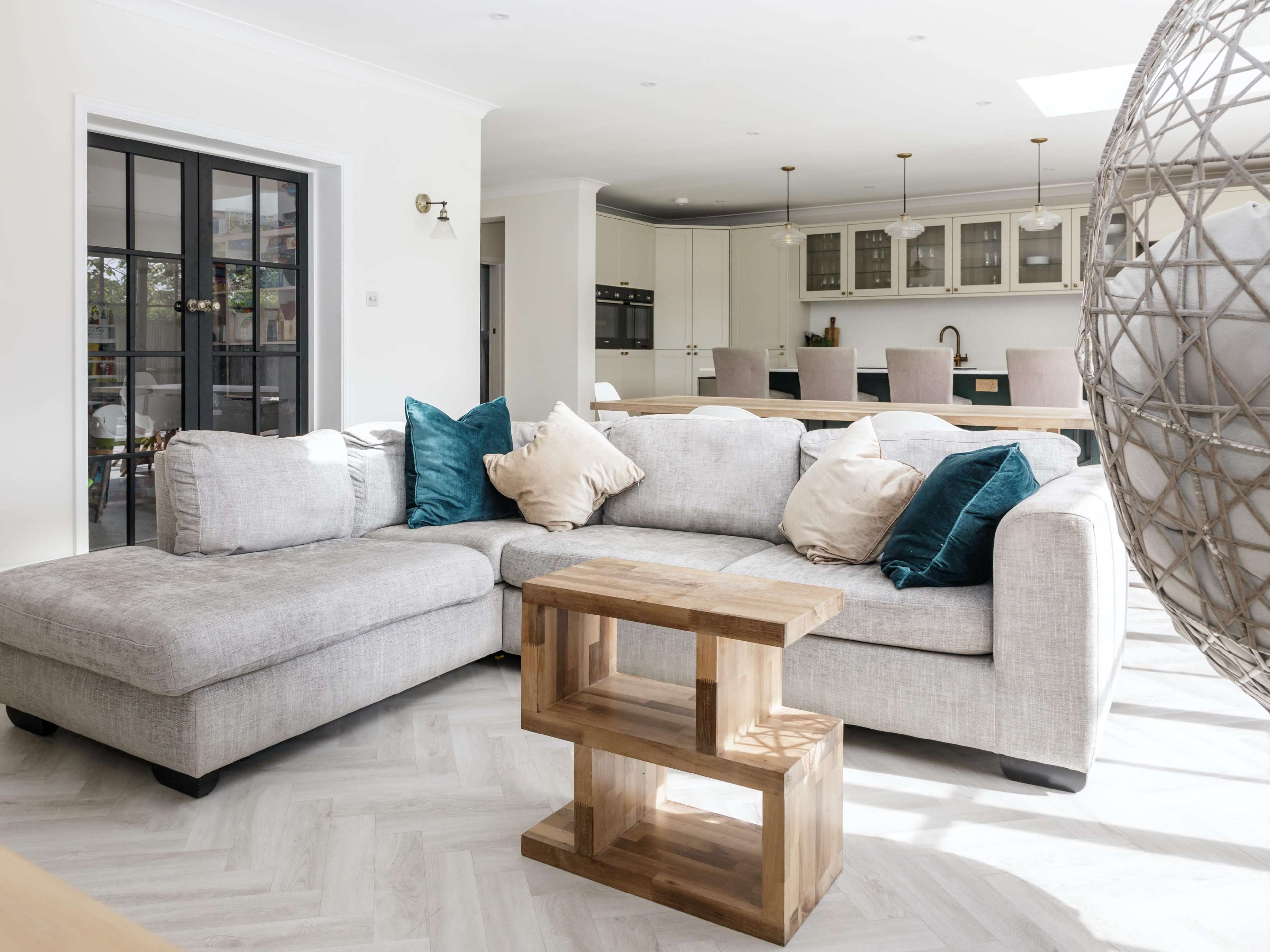 The image shows a modern living room featuring a light gray sectional sofa with decorative cushions and a wooden side table, set in an open-concept space that leads to a kitchen area.