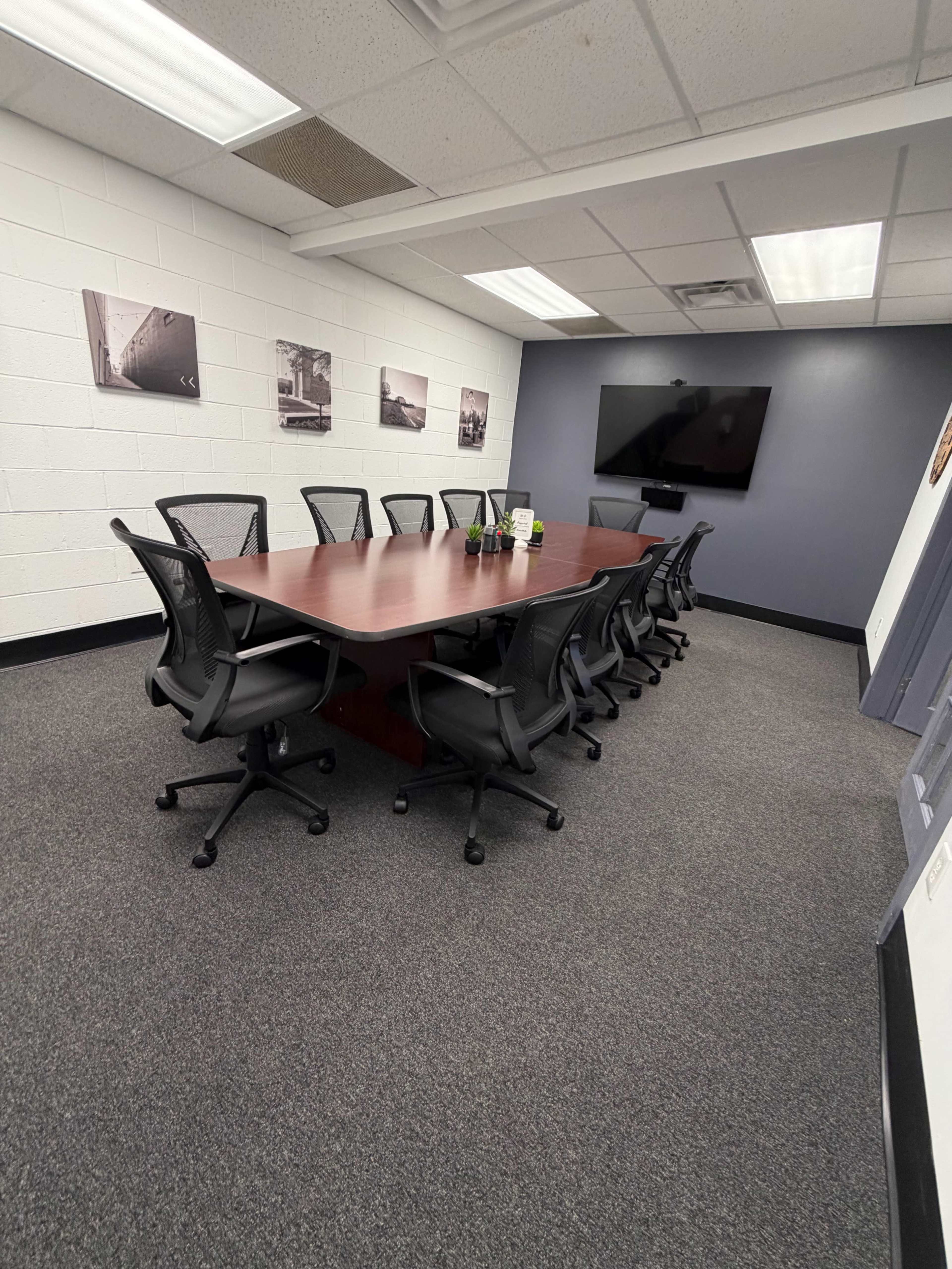 The image shows a conference room with a long rectangular wooden table surrounded by twelve ergonomic chairs, a television mounted on the wall, and several framed black-and-white photographs.