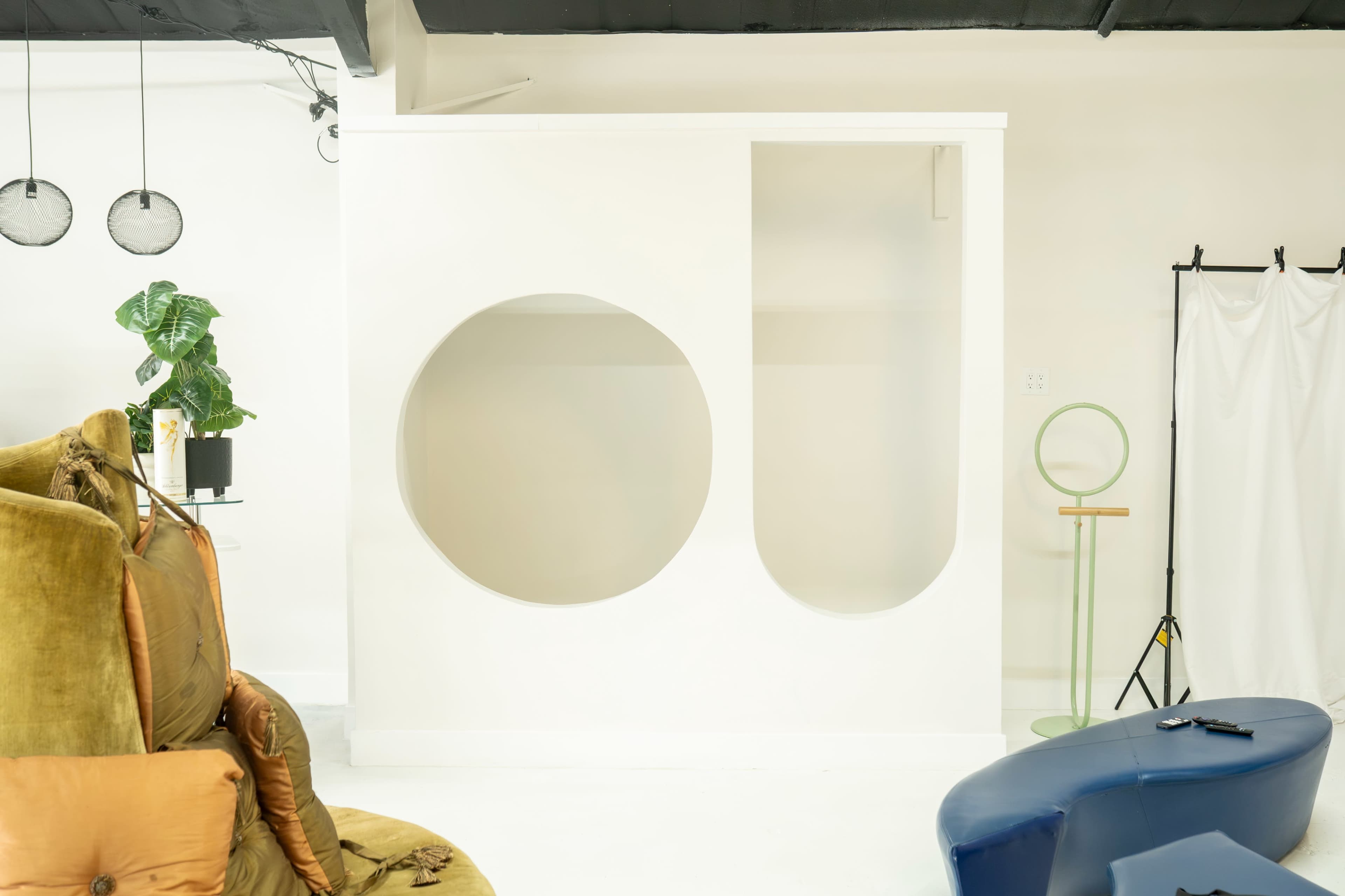 The image shows a minimalistic white wall partition with two circular cutouts, set in an interior space featuring a plant and various furniture pieces.