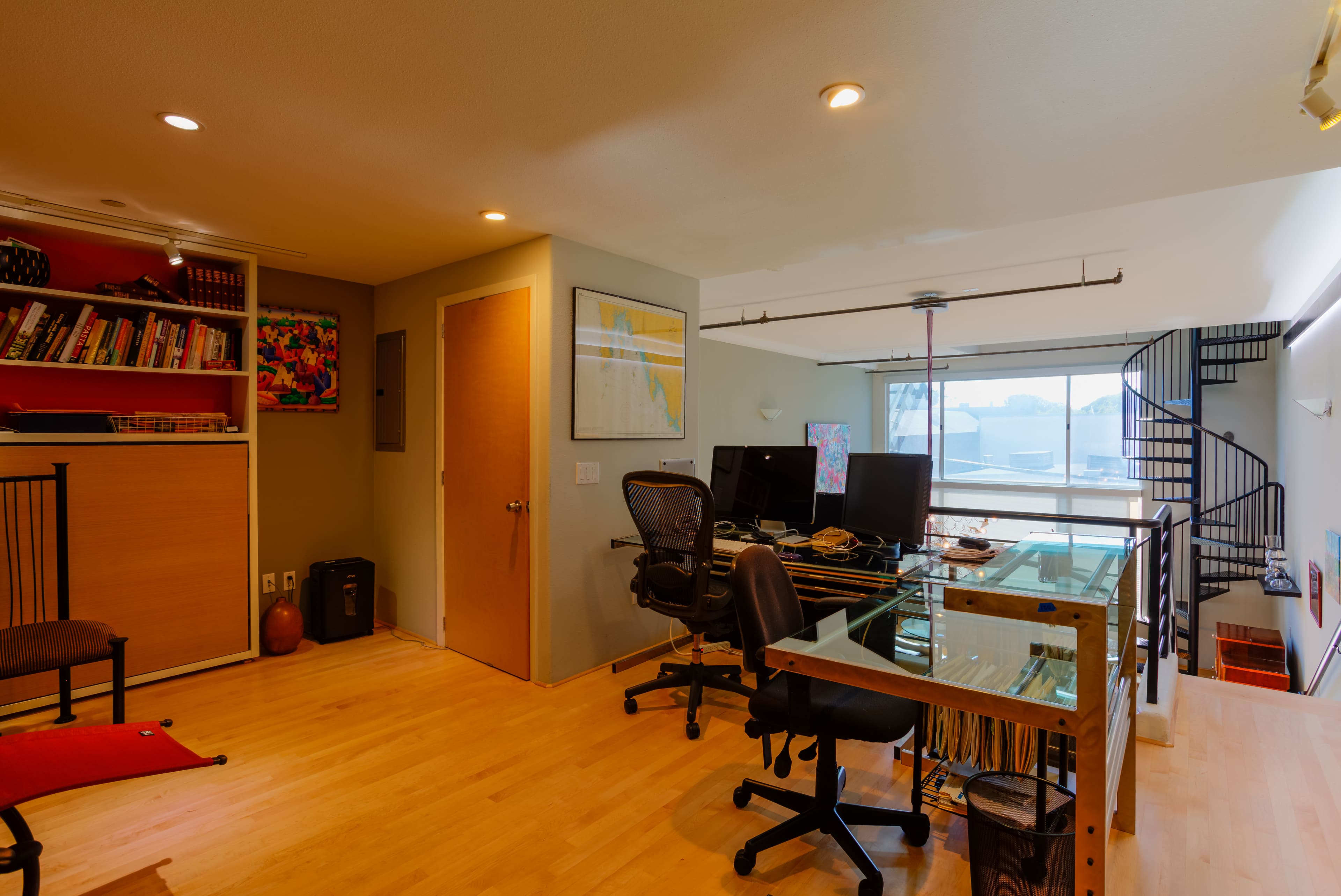 A modern office space with a glass desk, two computer monitors, and a staircase leading to an upper level.