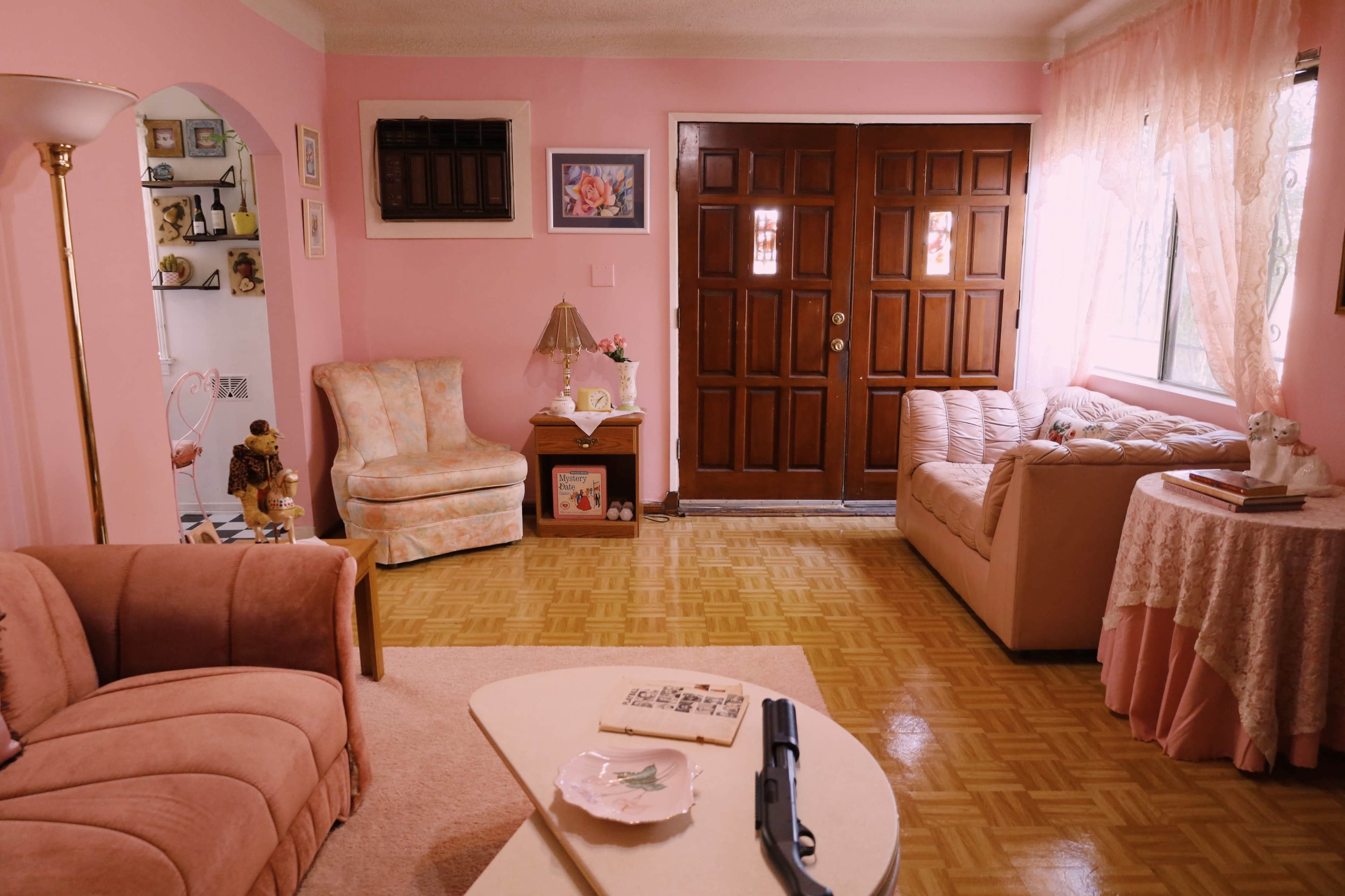 The living room features pink walls, a wooden double door entry, a beige armchair, a pink couch, and a coffee table with a decorative plate and a revolver on it.