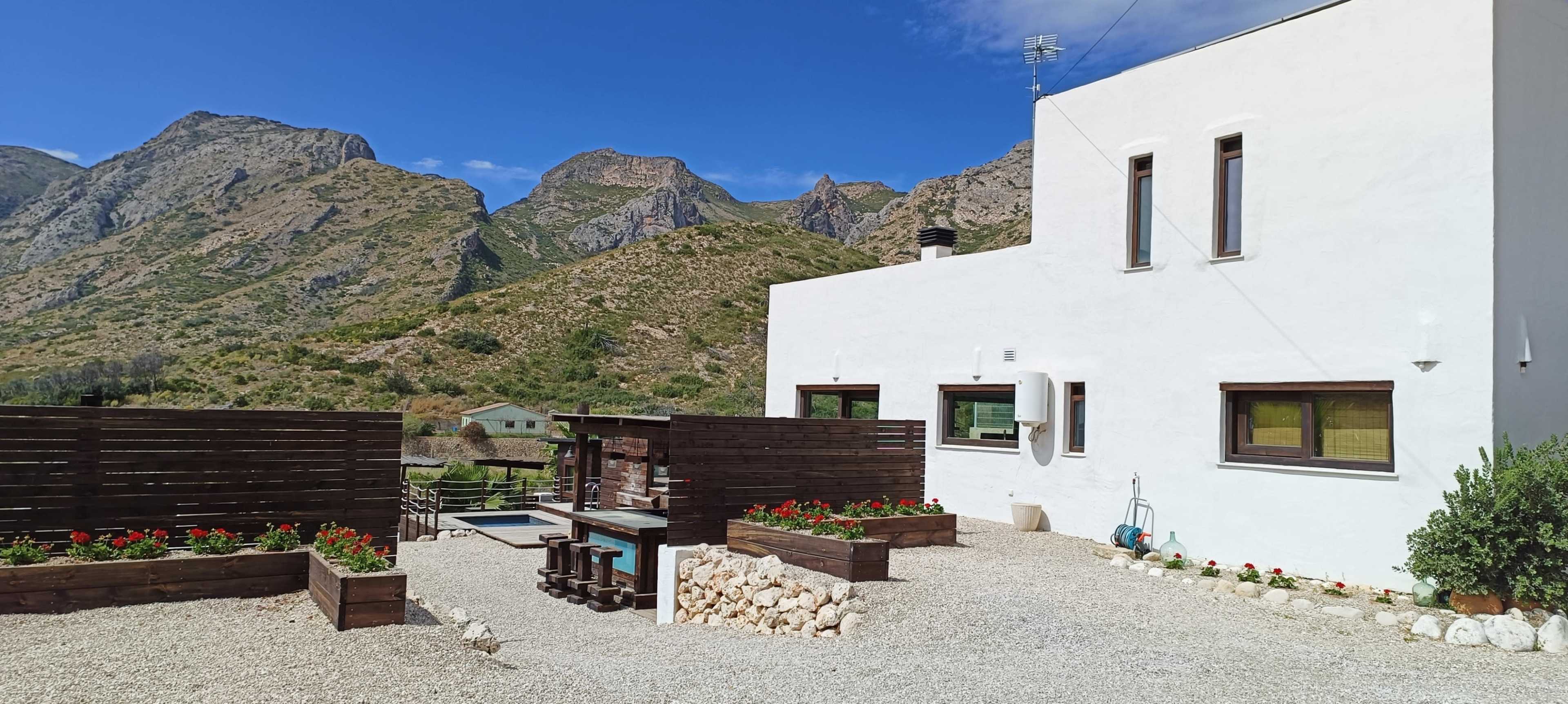 A modern white house with large windows overlooks a stone patio, flower beds, and a swimming pool, surrounded by mountainous terrain.
