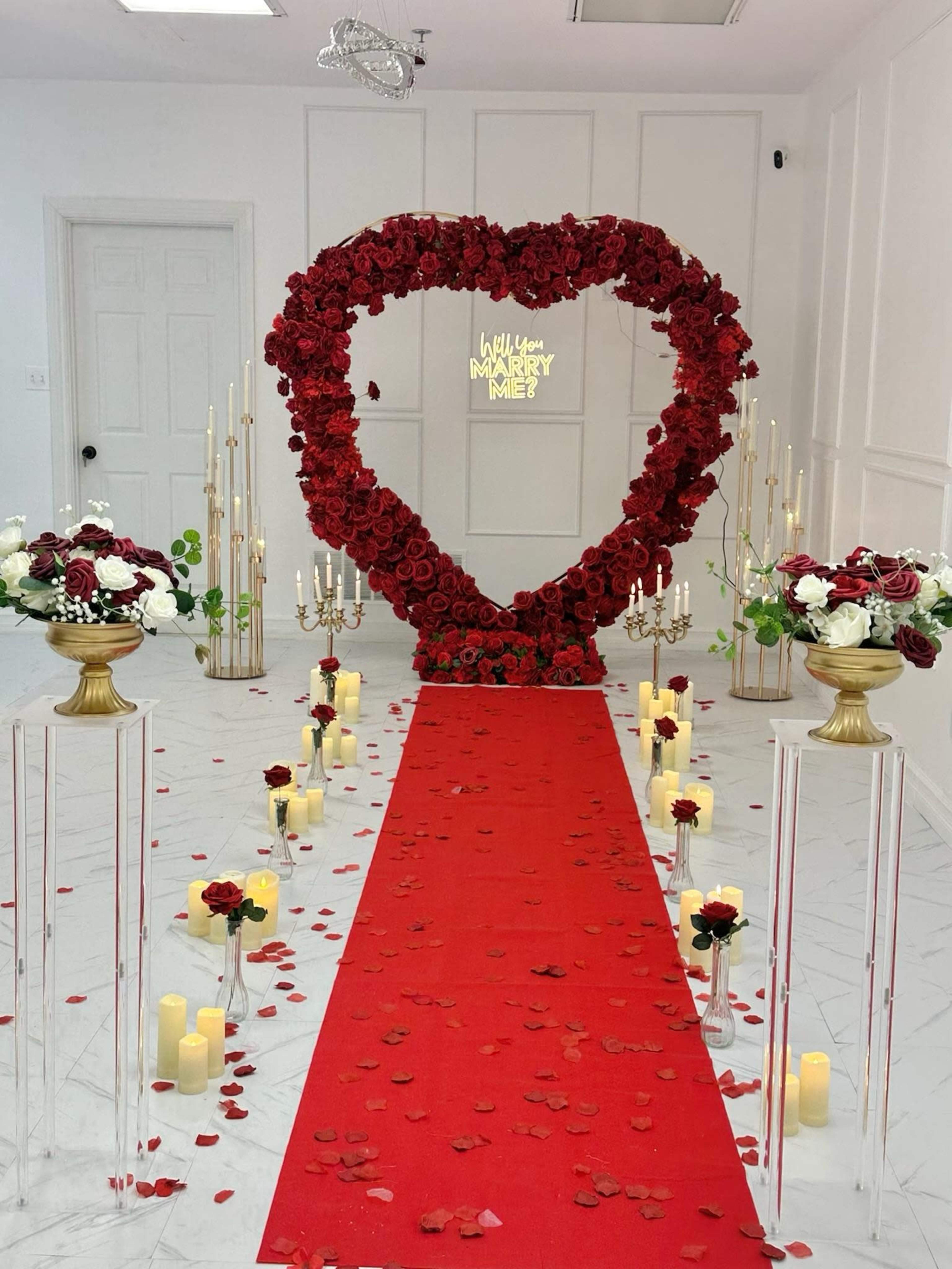 The image shows a decorative wedding setup with a large heart-shaped arrangement of red roses at the end of a red carpet, flanked by elegant centerpieces and candles along the aisle.