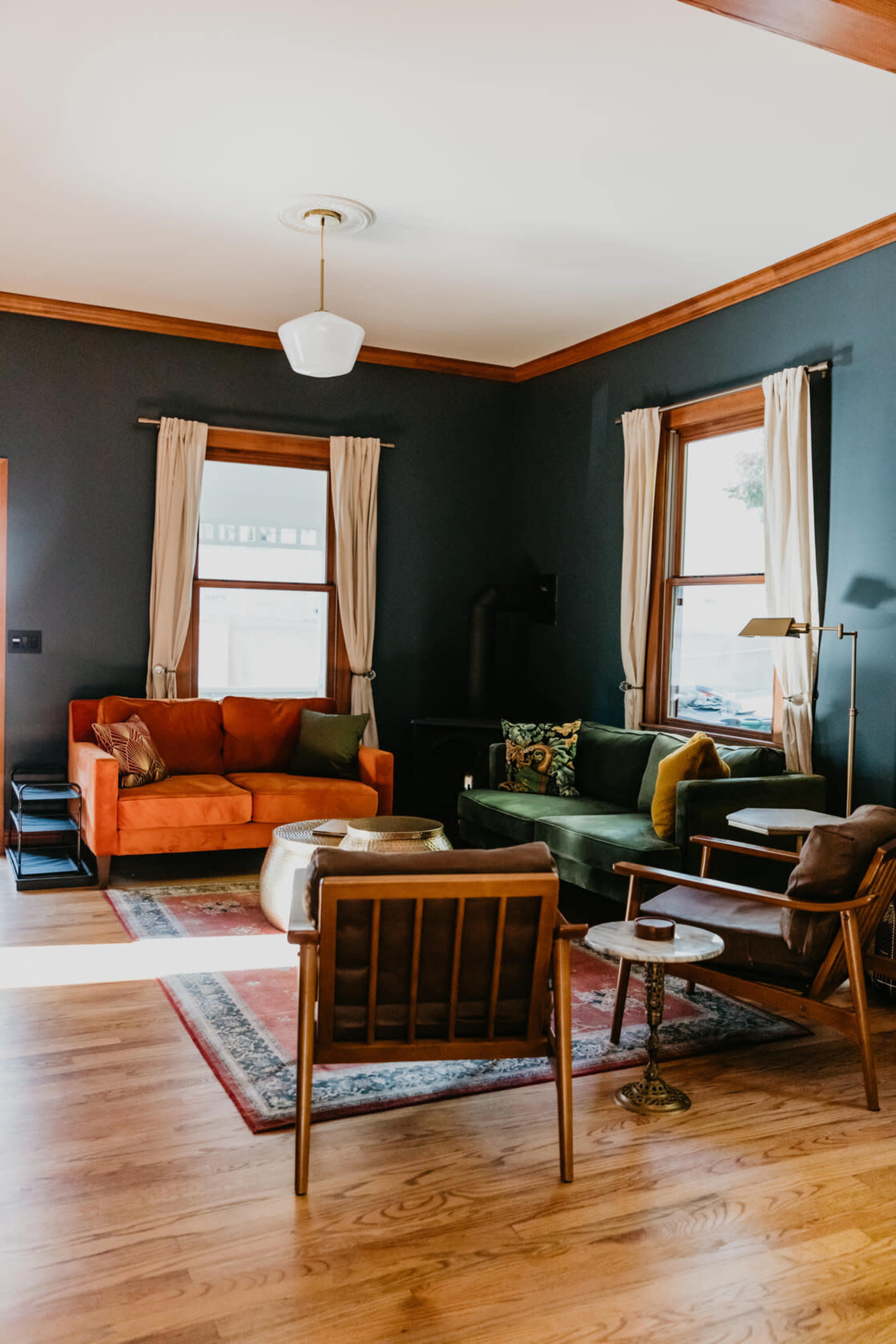 A living room with orange and green sofas, a wooden floor, and large windows framed by light curtains.