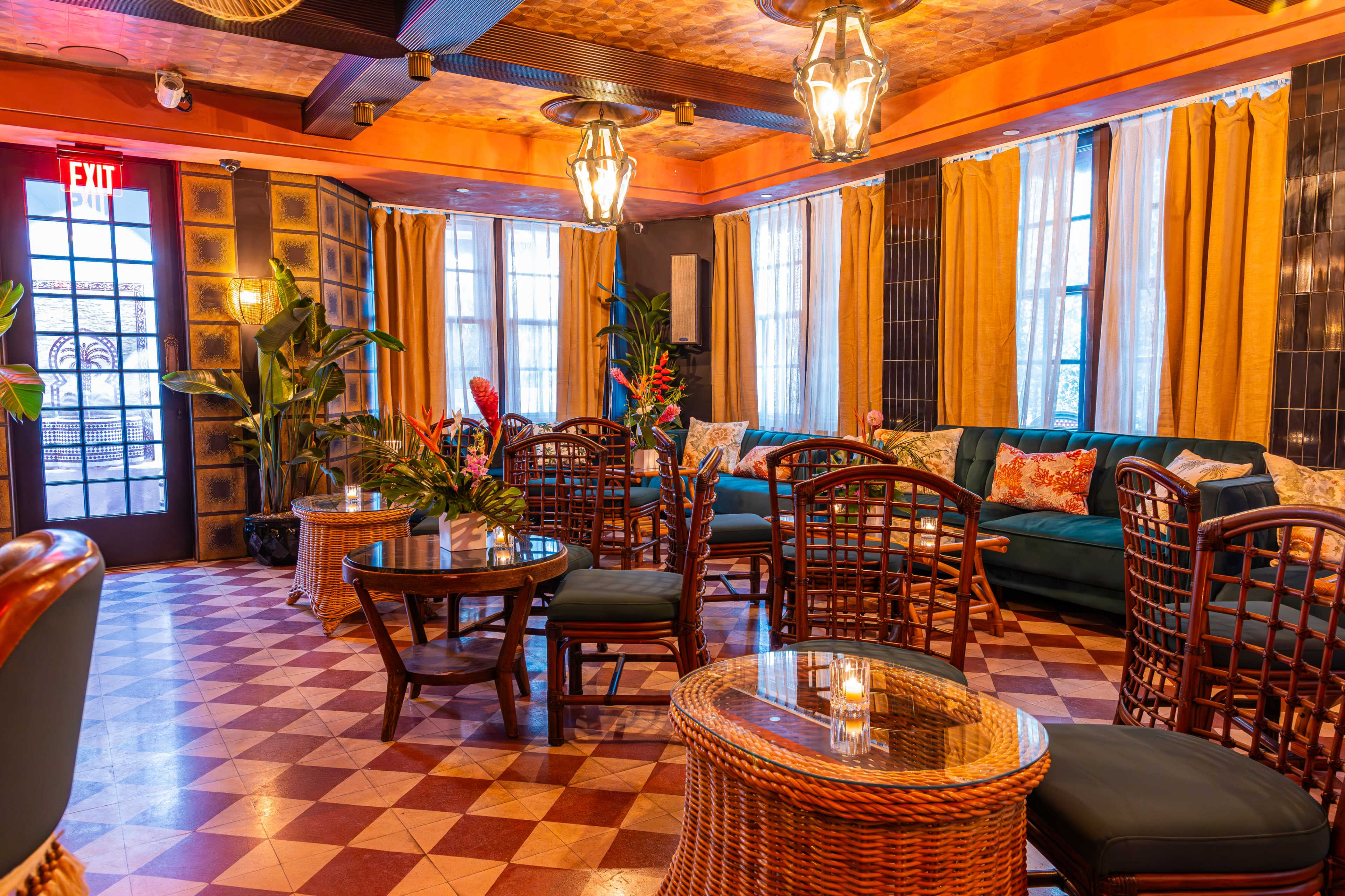 The image shows a cozy lounge area with rattan furniture, plants, and large windows allowing natural light to enter.